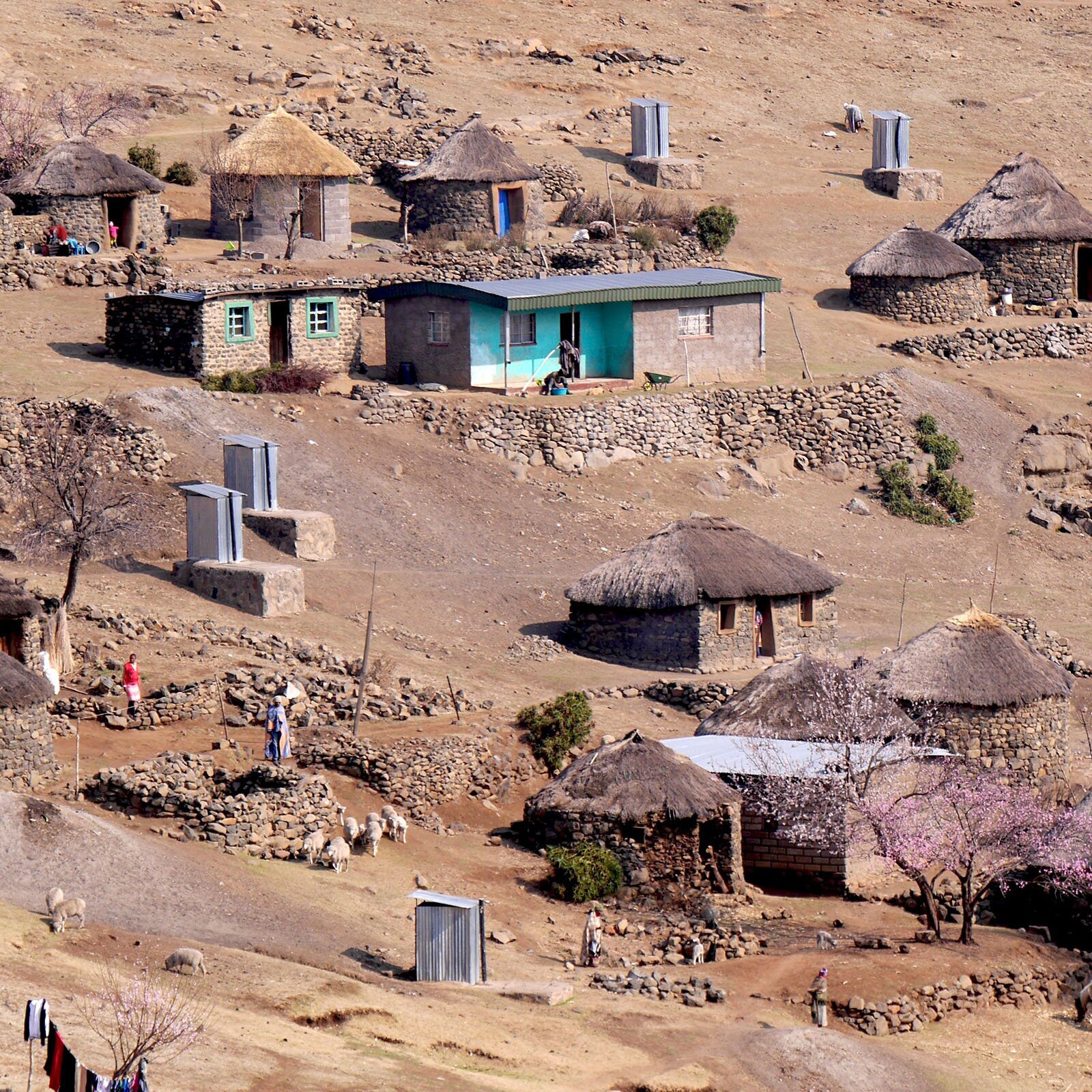 Zdjęcie przedstawia małą wioskę lub osadę, położoną na piaszczysto kamienistym terenie. Widzimy na nim małe domki. Większość zbudowana jest na planie okręgu i wzniesiona przy użyciu szarych i brązowych kamieni. Domki te pokryte są słomianą strzechą. Inne domki wybudowane są z szarej cegły na planie klasycznego prostokąta, posiadają okna i dwuspadowe, kryte dachy. Zdarzają się również domki stanowiące połączenie obu tych stylów. Przy domkach ustawione są małe, blaszane wychodki.