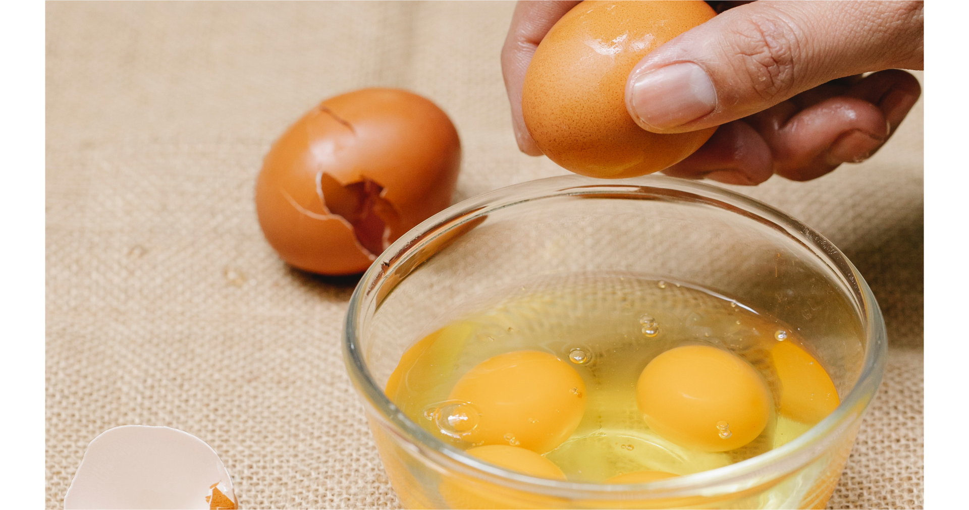 Zdjęcie przedstawia rękę osoby rozbijającej jajka do miseczki. W miseczce znajduje się kilka rozbitych już jajek, a obok miseczki widoczne są skorupki.