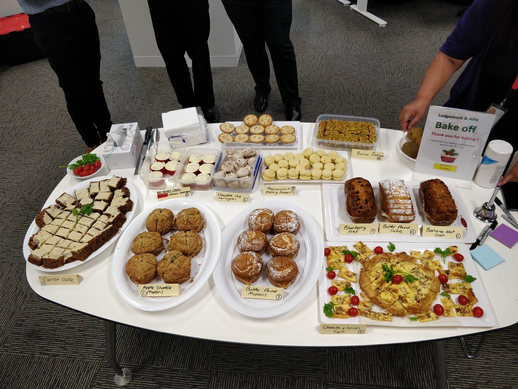 Zdjęcie przedstawia owalny stół z białym blatem, na którym widoczne są różnego kształtu i wielkości pojemniki oraz półmiski. Od lewej widoczny jest półmisek z kostkami ciemnego ciasta z białą polewą, przed którym stoi kartka z napisem w języku angielskim Carrot cake i numer 1. Na prawo widoczny jest półmisek z babeczkami, na którym stoi kartka z napisem w języku angielskim Apple Crumble Muffins i numer 2. Na prawo widoczny jest półmisek z babeczkami, na którym stoi kartka z napisem w języku angielskim Butter Pound Muffins i numer 3. Na prawo widoczny jest półmisek z okrągłym ciastem pokrojonym na części oraz fragmentami ciasta. Na półmisku stoi kartka z napisem w języku angielskim Cheese &amp; onion Tart i numer 4. Powyżej widoczny jest półmisek z wypiekami, a którym widać trzy kartki z napisami w języku angielskim Blueberry loaf i numer 9, Butter Pound Cake i numer 10, Banana Nut loaf i numer 10. W górnej części stołu widoczne pojemniki z wypiekami z kartkami z nieczytelnymi napisami w języku angielskim. W prawym górnym rogu blatu widoczna jest kartka z napisem w języku angielskim Lodgement &amp; APls Bake off Thank you for baking! oraz rysunkiem babeczki. Przy blacie stoją cztery osoby, widoczne od pasa w dół.