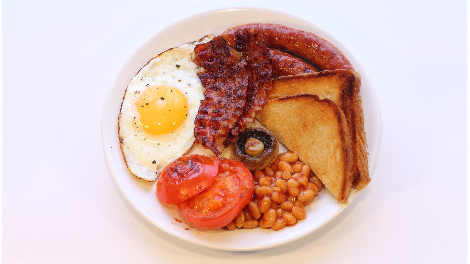 Zdjęcie przedstawia talerz, na którym ułożono jajko sadzone, tosty, pieczony bekon, pomidor smażony , pieczarkę i fasolę w sosie pomidorowym.