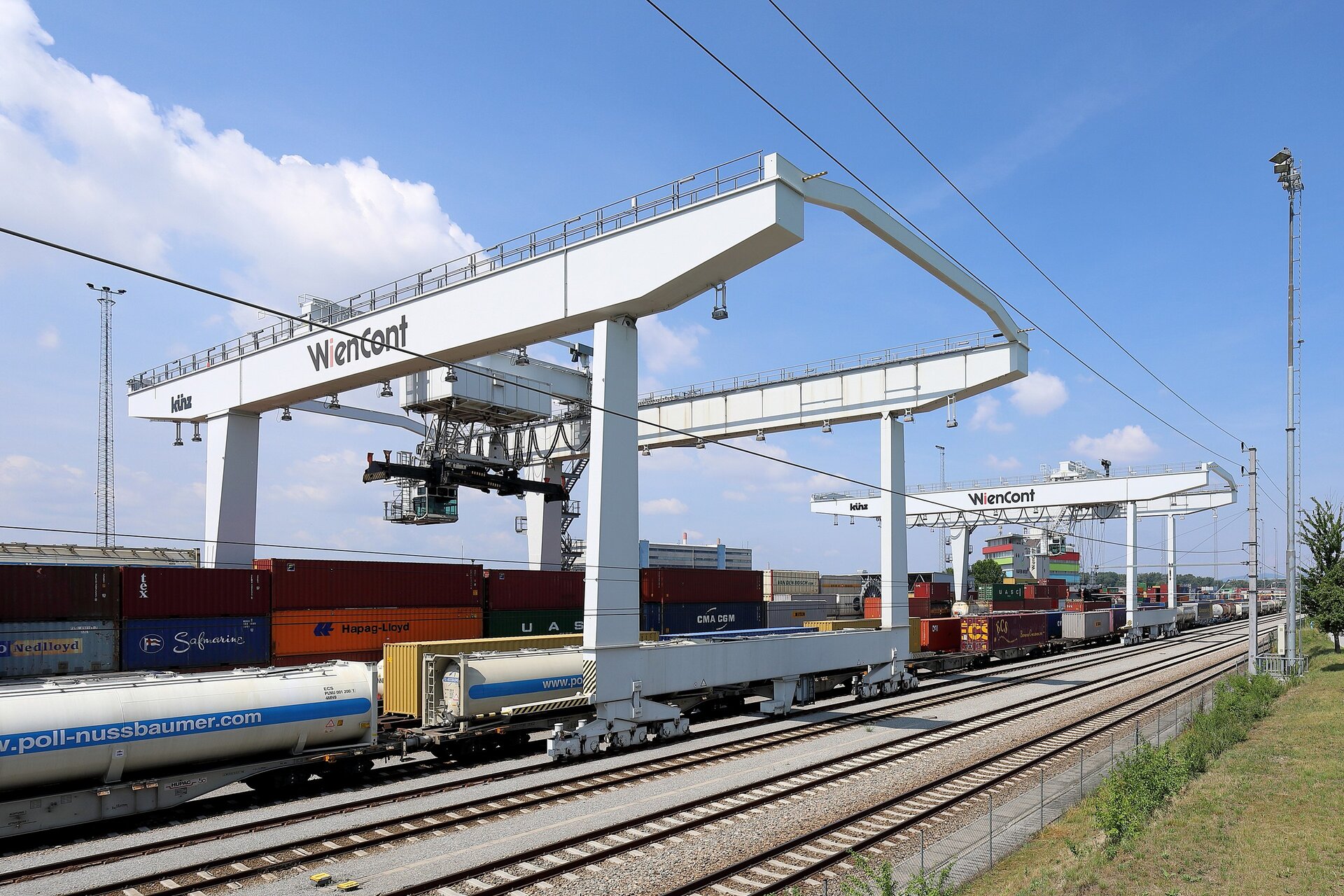 Zdjęcie przedstawia torowisko na terenie otwartym .Widoczne są liczne cysterny i kontenery. Ponad nimi stoi stalowa konstrukcja. To dwa połączone moduły ram z podczepioną kabiną operatora i podwieszonymi wyciągami oraz systemem lin.