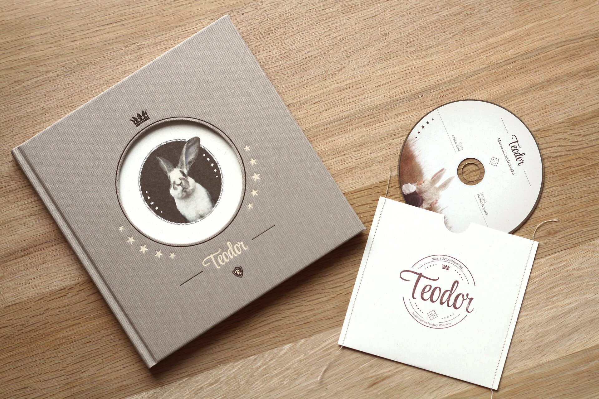 Ilustracja przedstawia okładkę książki dla dzieci Marii Szczodrowskiej: „Teodor”. Na drewnianej powierzchni leży zamknięta książka oraz płyta CD. W okrągłym okienku, imitującym króliczą norę znajduje się biały królik o lekko szarawych uszach i pyszczku. Pod okienkiem biały napis: Teodor, po obu stronach białe gwiazdki. Tło okładki jest jasnobrązowe. Z prawej strony leży białe, papierowe opakowanie płyty CD z brązowym napisem na środku: Teodor. Z opakowania wysuwa się płyta CD, na której znajduje się napis: Teodor, pod nim dwoje ludzi na łące, opartych o siebie plecami, na głowach mają peruki w kształcie króliczych uszu. Po bokach napisy: Czyta Olga Bołądź, muzyka Michał Jacaszek.