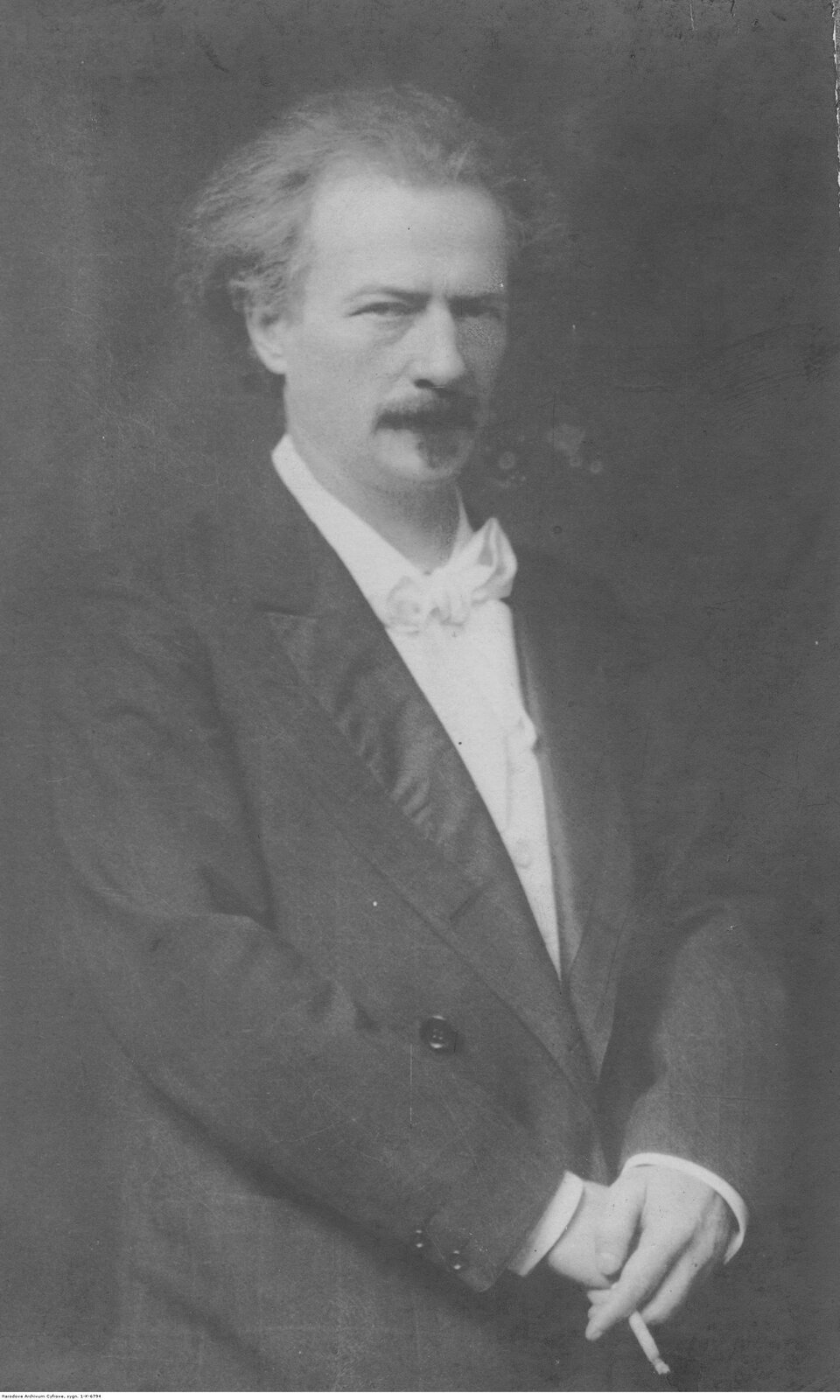 Ilustracja przedstawia Ignacego Jana Paderewskiego. Mężczyzna w średnim wieku z dużymi zakolami. Ma dłuższe, rzadkie włosy oraz brodę i wąsa. Ma zadziorny i prowokujący wyraz twarzy. Ubrany jest w białą koszulę i marynarkę. Ręce ma skrzyżowane w pasie, w lewej ręce trzyma odpalonego papierosa.