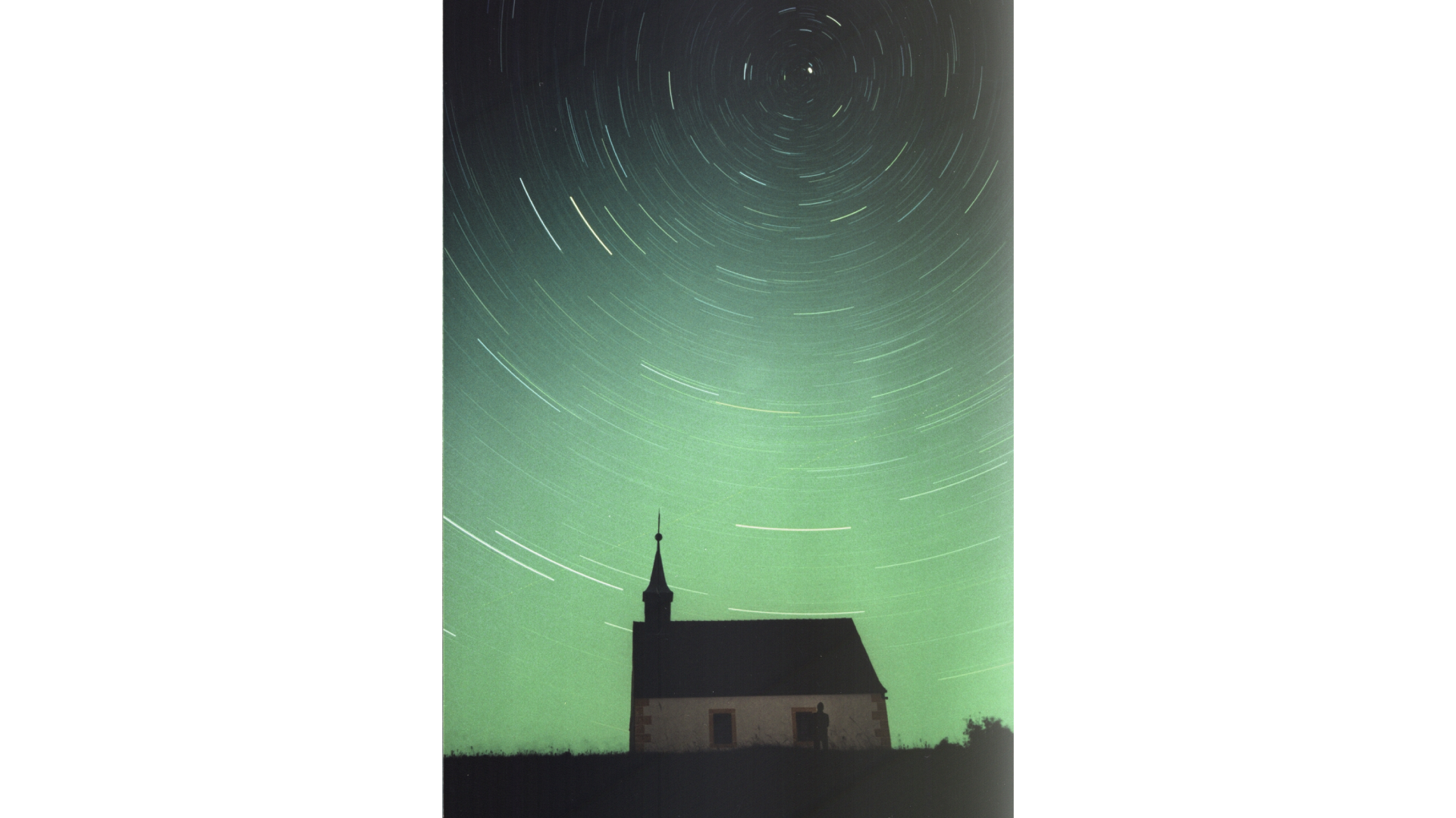 Zdjęcie przedstawia tory gwiazd okołobiegunowych na wieczornym niebie. Tworzą one koncentryczne kręgi wokół punktu, w którego pobliżu widać jaśniejszą kropkę, czyli Gwiazdę Polarną. Na pierwszym planie fotografii widać mały kościół.