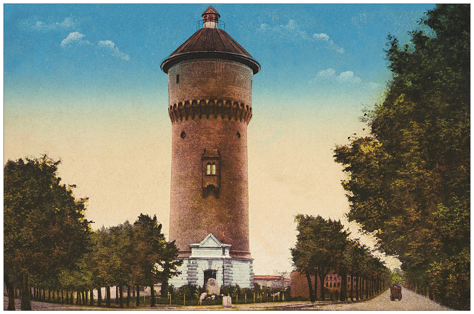 Zdjęcie przedstawia wygląd wieży ciśnień zbudowanej na planie koła. Budowla zbudowana jest z cegły w stylu neoromańskim z hełmem. Posiada kamienną obudowę podstawy i wejście z trójkątnym przyczółkiem. Znajduje się na rogu zbiegu ulic, wzdłuż których rosną równe, zielone drzewa.