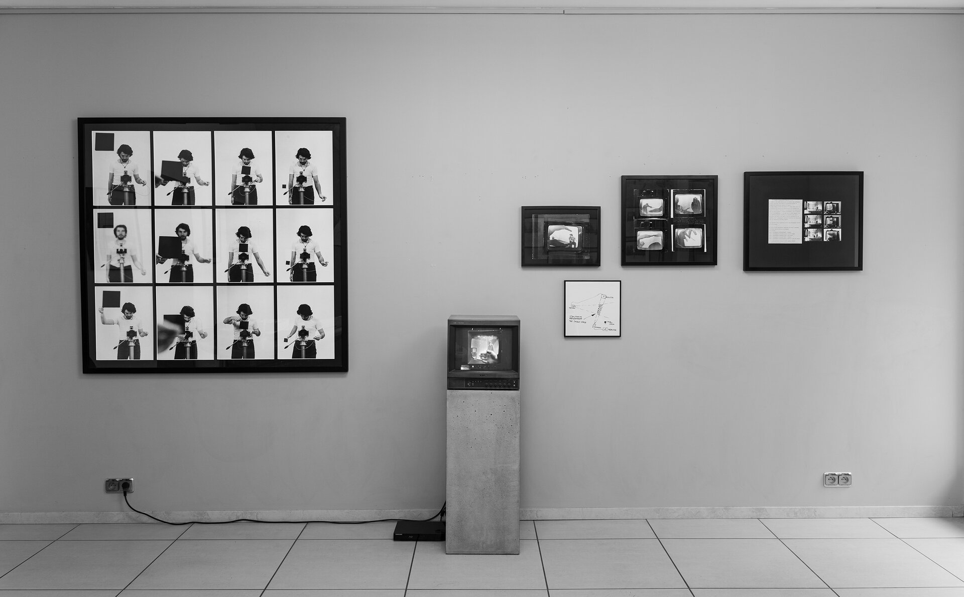 Na fotografii ukazano pomieszczenie. Na ścianie po lewej stronie znajduje się kolaż złożony z dwunastu zdjęć, na których widnieje mężczyzna robiący zdjęcia. Po środku fotografii znajduje się kolumna z małym telewizorem. Po prawej stronie znajdują się cztery obrazy. Jeden z nich tworzy kolaż czterech prac. Fotografia jest czarno‑biała. 