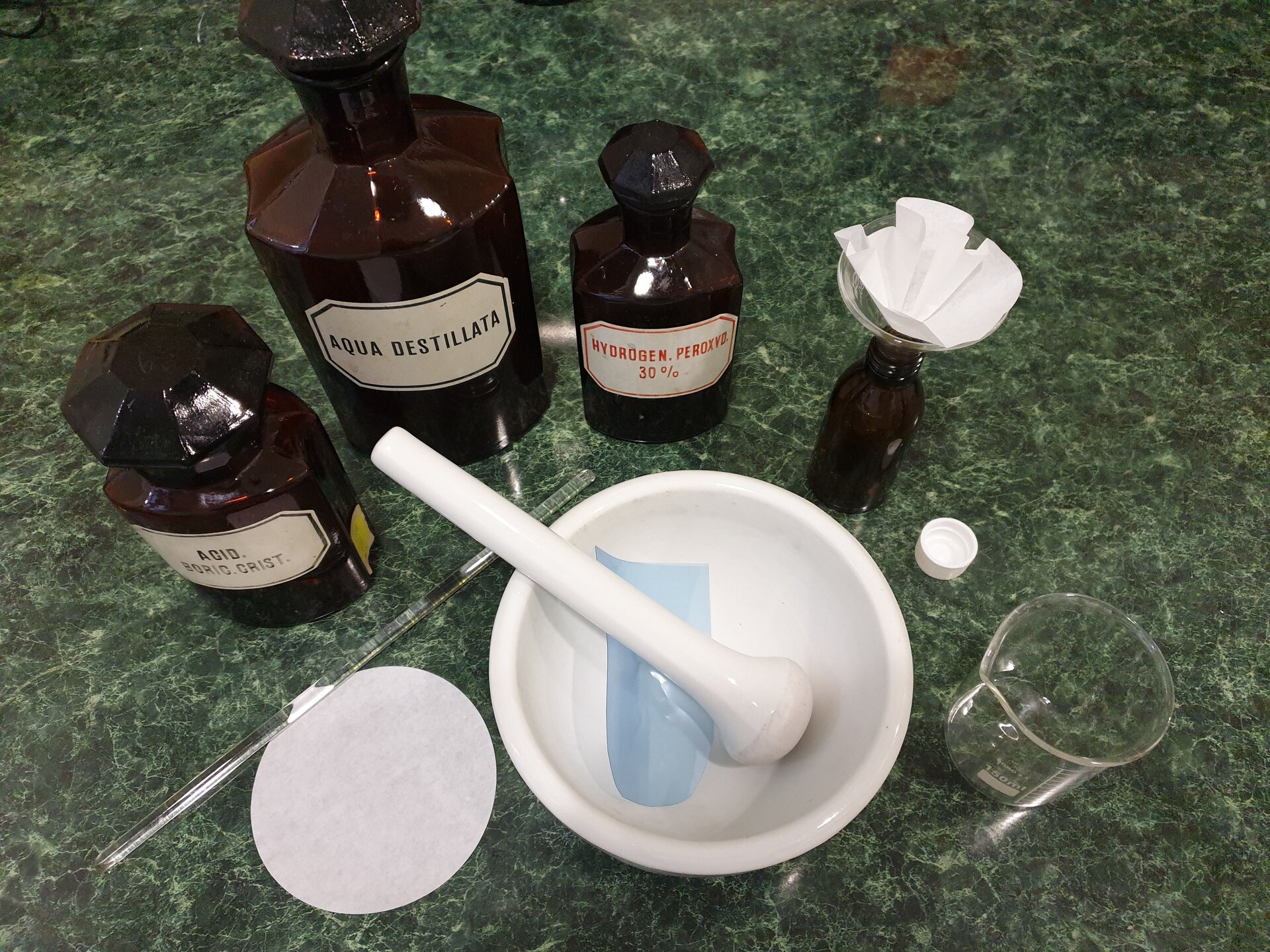 Rycina 1 przedstawia stojące obok siebie szklane butelki ze szlifem, czyli ze szklanym dopasowanym zamknięciem w formie stożkowatej. Każda posiada podpisaną etykietę. Butelki mają różne rozmiary. Obok znajduje się moździerz, niewielka zlewka, sączek oraz szklane mieszadełko w postaci patyczka. Obok stoi otwarta butelka, w którą wetknięto lejek. W lejek włożono sączek, czyli okrągłą bibułkę filtracyjną.