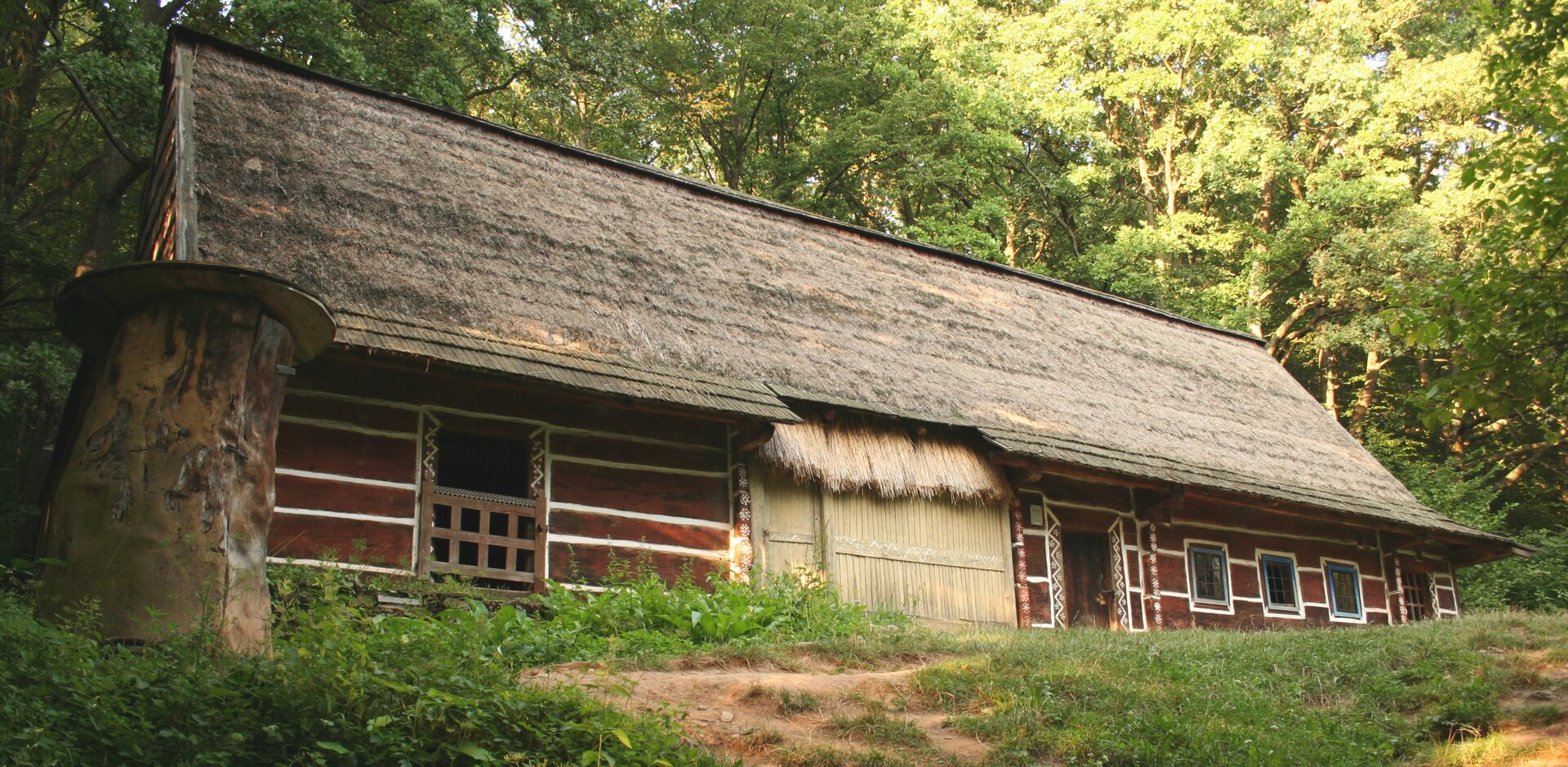 N zdjęciu podłużny, drewniany dom. Ma spadzisty dach, kryty strzechą. Białe zdobienia na łączeniach belek i wokół okien.