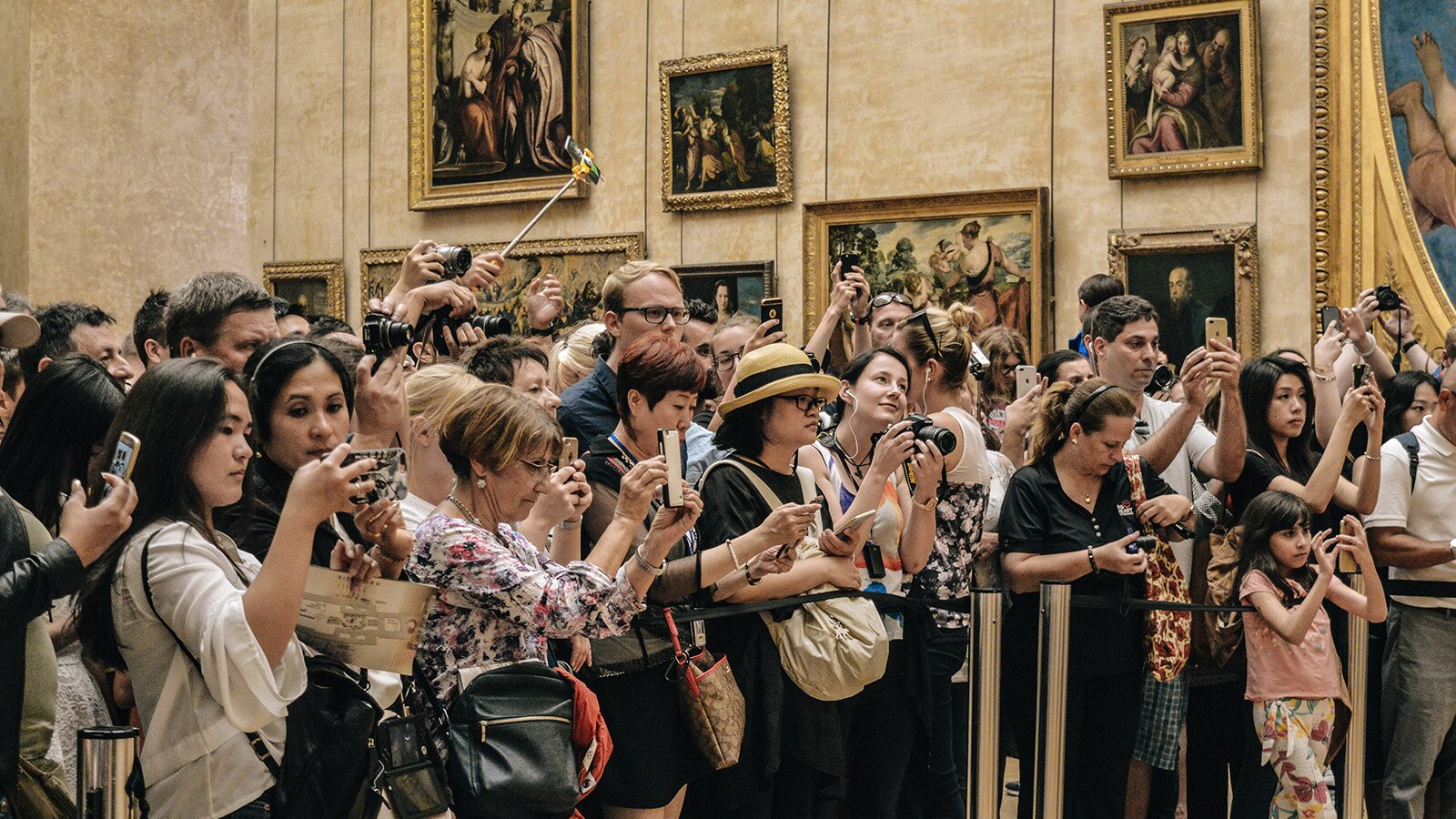 Zdjęcie przedstawia wnętrze galerii sztuki. Na ścianach wiszą obrazy w złotych ramach przedstawiające sceny rodzajowe. Na pierwszym planie widoczny jest tłum ludzi. Widoczne są osoby  trzymające w dłoniach telefony i robiące zdjęcia. Są tez osoby z aparatami fotograficznymi. Ktoś w tłumie trzyma telefon na długim statywie. 