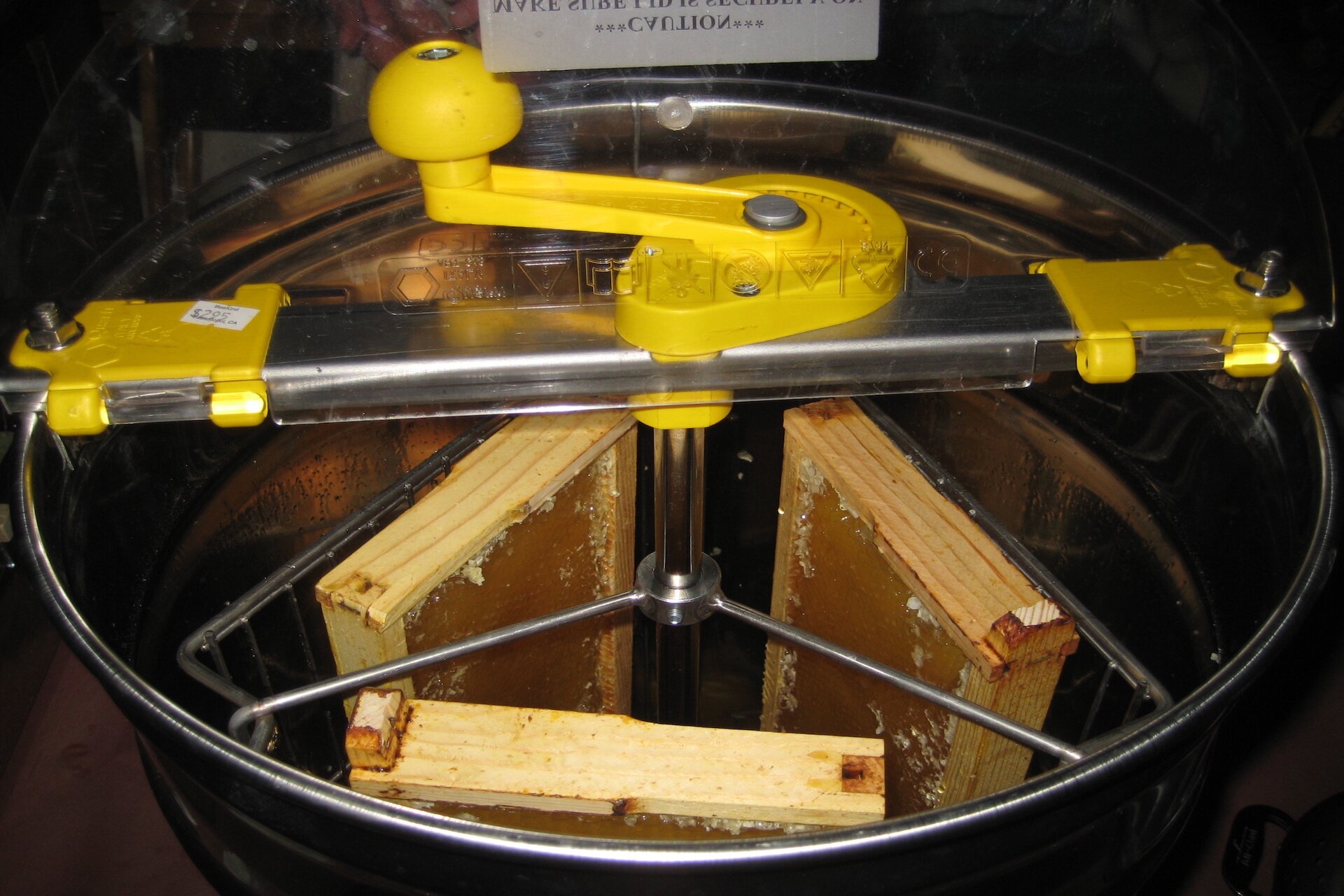 Zdjęcie przedstawia miodarkę. Jest to duży pojemnik w kształcie walca z metalowym koszyczkiem, do którego włożone są plastry pokryte miodem.