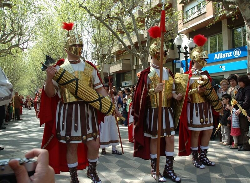 Zdjęcie przedstawia trzech mężczyzn w strojach rzymskich legionistów. Mężczyźni idą ulicą miasta. Po obu stronach ulicy widać stojący tłum ludzi we współczesnych strojach.