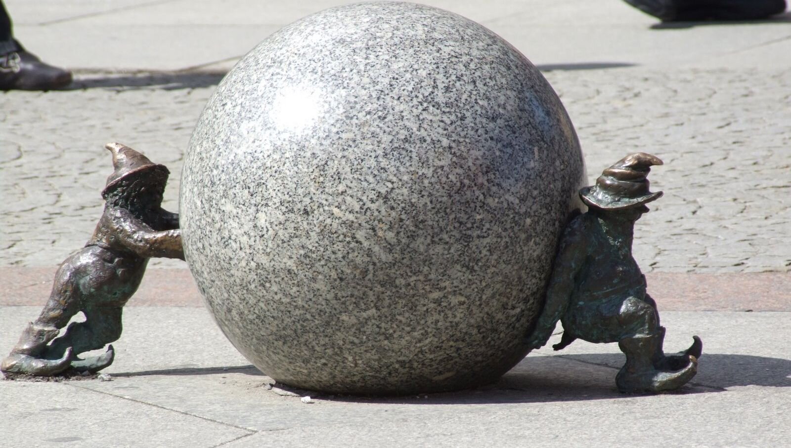 Kliknij, żeby powiększyć. Zdjęcie rzeźby przedstawiającej wielką, kamienną kulę, którą próbują przesunąć dwa małe krasnale. Jeden ją pcha, a drugi blokuje kontrolując tempo toczenia.