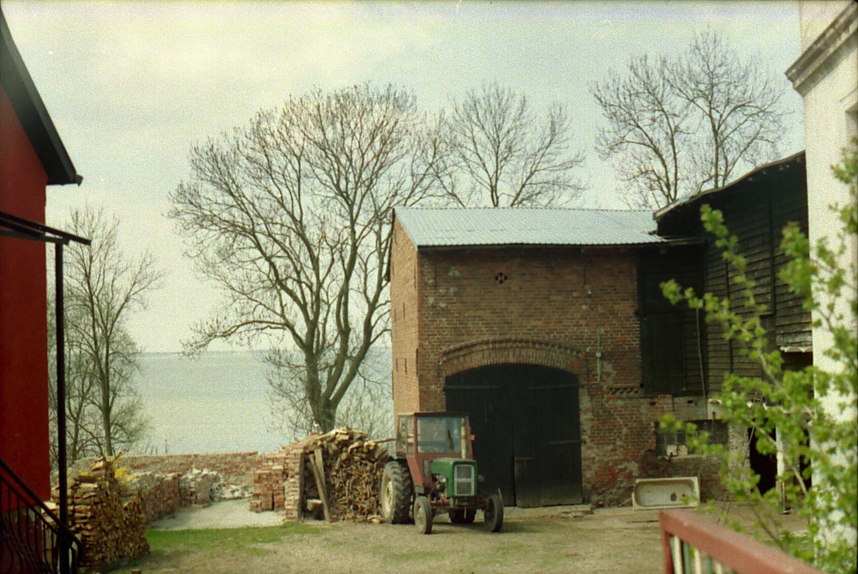 Kliknij, aby powiększyć. Zdjęcie przedstawia fragment gospodarstwa. Na pierwszym planie po lewej fragment domu. Po prawej fragment innego domu, dalej chlew i traktor w środku podwórka. W tle między budynkami jezioro.