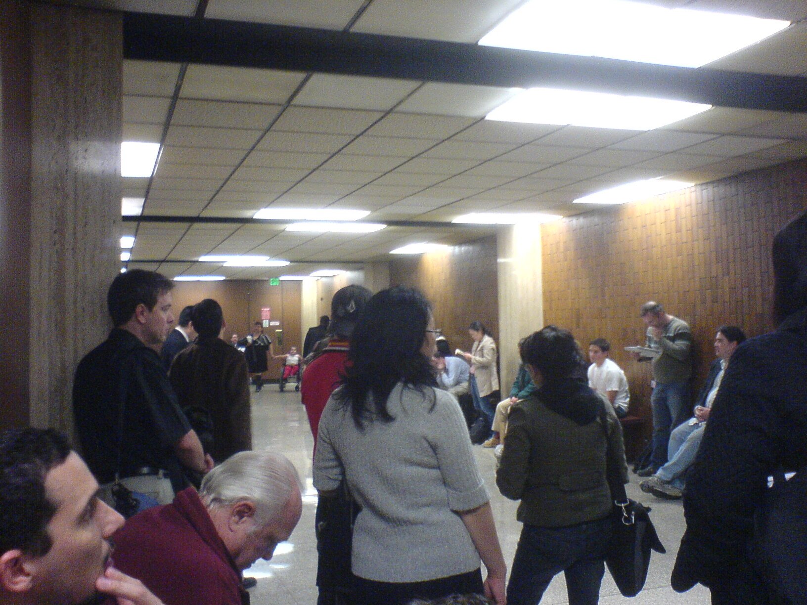 Zdjęcie przedstawia korytarz, w którym stoją i siedzą licznie zgromadzeni ludzie.