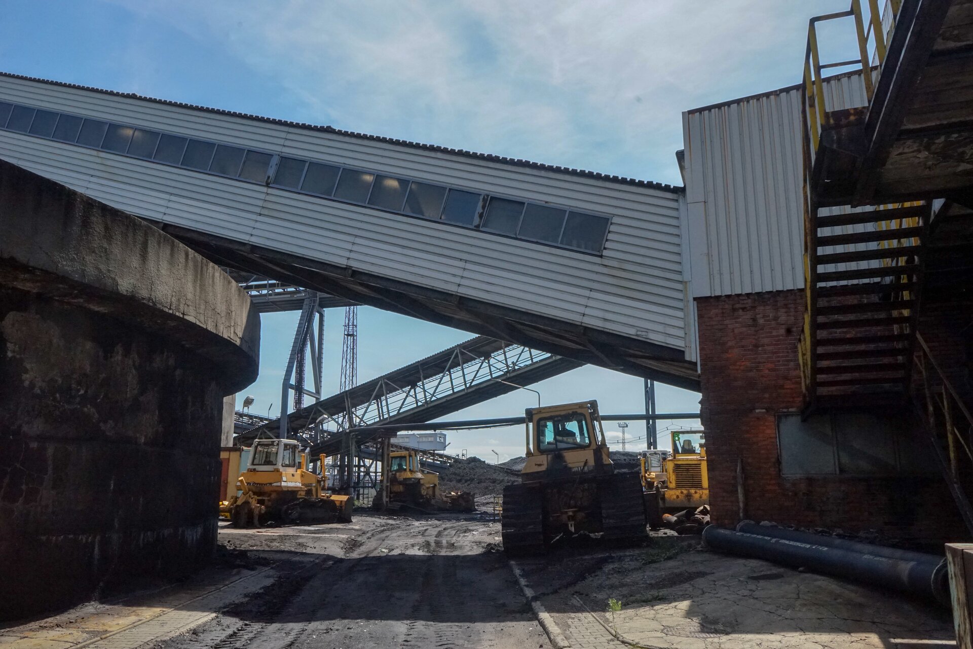 Na zdjęciu widoczny jest teren przemysłowy widoczny z poziomu gruntu. W centralnej części zdjęcia znajduje się most transportowy łączący dwa budynki widoczne po obu stronach zdjęcia. Łącznik i budynki wykonane są z blachy falistej. Łącznik osadzony jest na stalowych nogach. Pod mostami znajduje się droga przemysłowa. Po drodze poruszają się cztery duże spychacze gąsienicowe. W tle widać kolejny most. Obok niego znajduje się czarna hałda węgla. Poniżej zdjęcia widoczny jest przycisk multimedialny z nagraniem audio. Treść nagrania jest zgodna z tytułem zdjęcia.