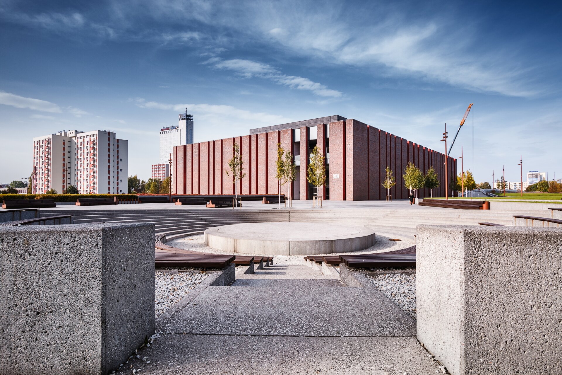 Zdjęcie przedstawia budynek Narodowej Orkiestry Symfonicznej Polskiego Radia w Katowicach. Na pierwszym planie zdjęcia ukazane są drewniane ławki. I szary murek. W oddali znajdują się drzewa oraz czerwony budynek.  Po lewej stronie znajduje się osiedle z wysokimi blokami. 