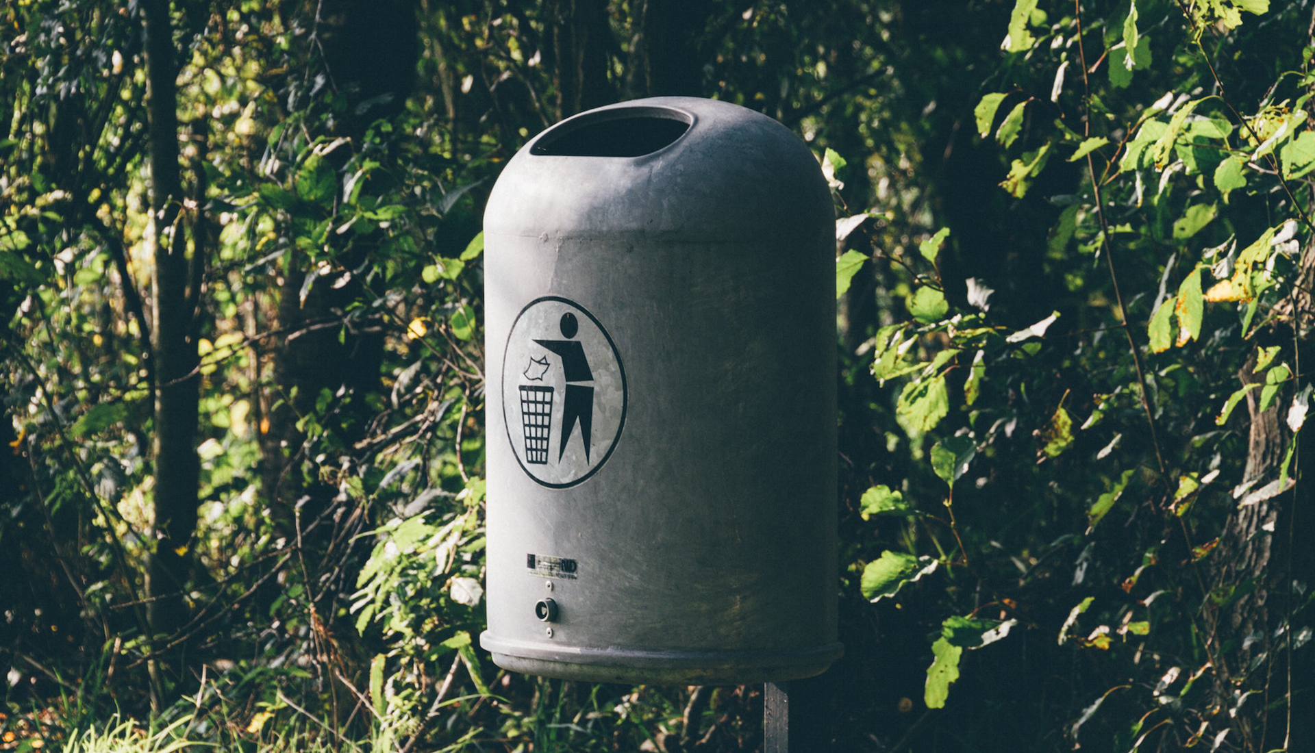 Zdjęcie przedstawia metalowy, okrągły kosz na śmieci. Znajduje sie na nim nalepka przedstawiająca postać wrzucającą śmieć do kosza. 