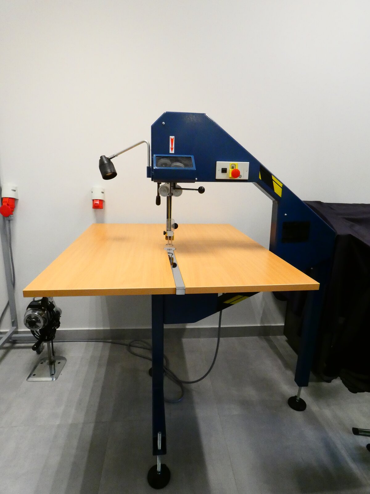 Zdjęcie przedstawia granatowe ramię maszyny zwisające nad drewnianym blatem. Z ramienia wychodzi igła, są na nim umieszczone przyciski, a także lampa, oświetlająca stanowisko pracy.