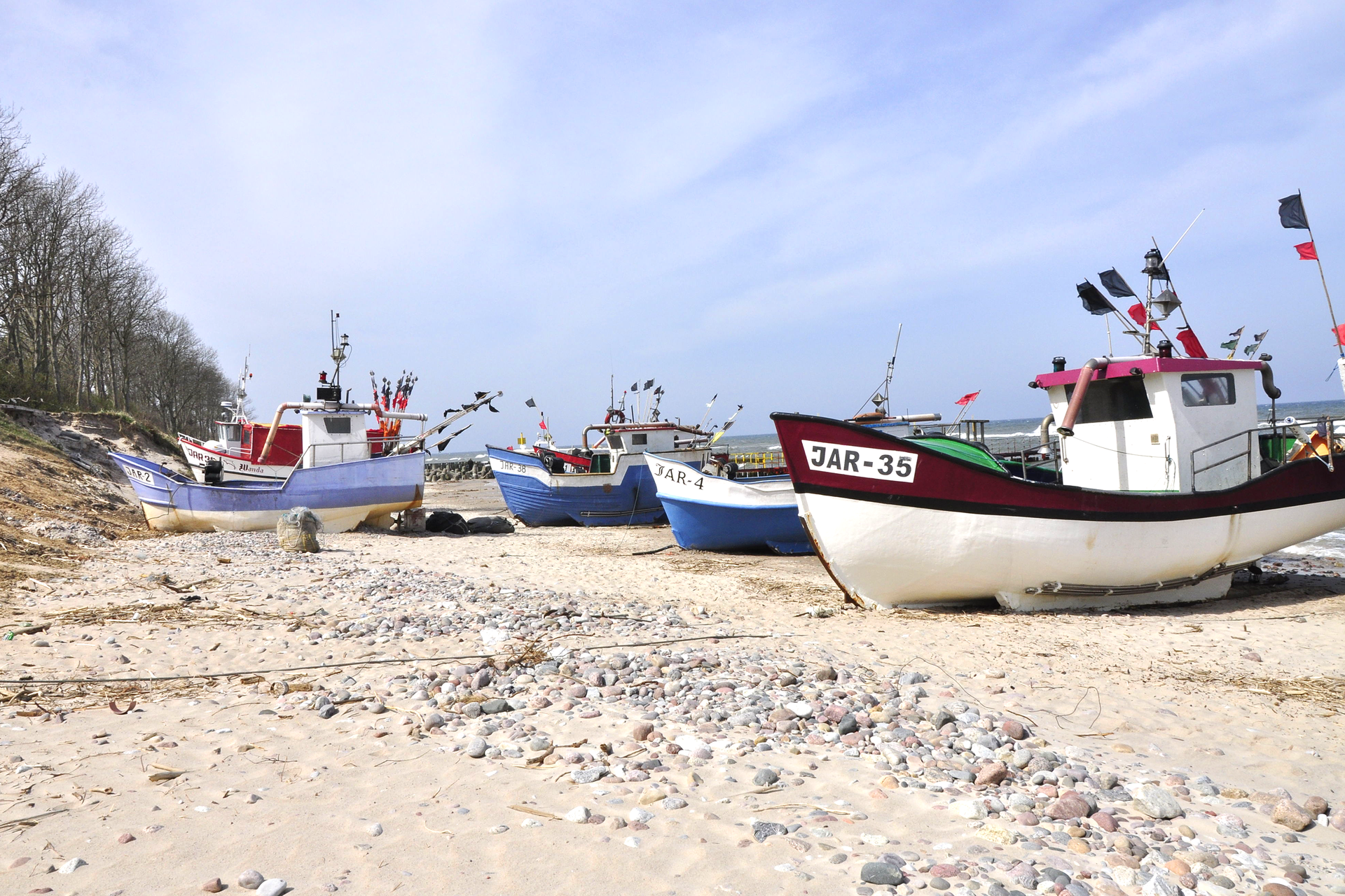 Fotografia przedstawiająca kutry rybackie na brzegu morza. W tle fotografii widać morze oraz niebo stykające się z morzem na horyzoncie. Przed kutrami, po lewej stronie zdjęcia, znajduje się niewysoki klif porośnięty drzewami liściastymi. Na pierwszym planie plaża z licznymi drobnymi kamieniami.