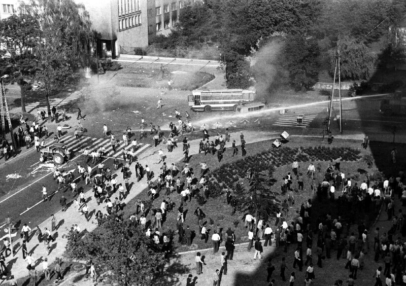 Czarno-biała fotografia przedstawiająca starcia manifestantów z milicją na ulicach Radomia w 1976 roku. Milicja używa armatek wodnych. Zdjęcie wykonano z lotu ptaka.