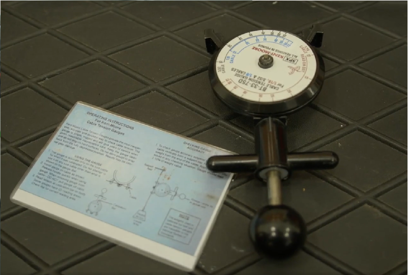 Zdjęcie przedstawia tensometr analogowy. Na podłożu leży urządzenie z okrągłą tarczą zegarową. Z niej wystaje z jednej strony cylinder z dwoma rączkami na końcu, w którym umieszczony jest pręt zakończony czarną kulką. Obok tensometru leży jego instrukcja użytkowania.