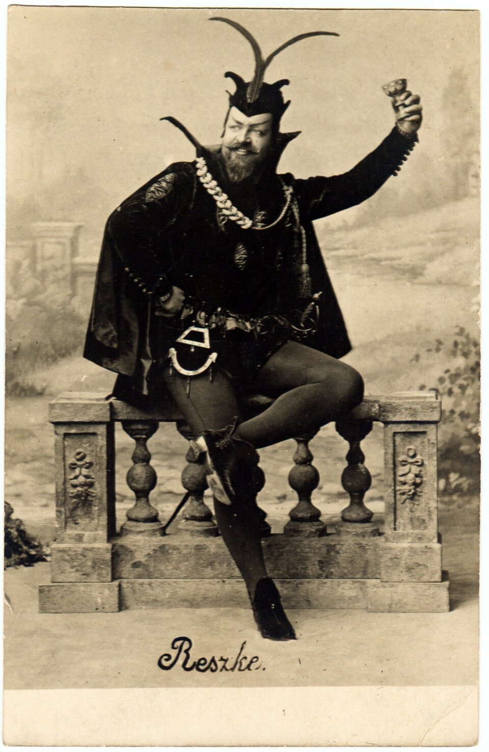 Fotografia przedstawia mężczyznę w sile wieku. Postać ukazuje lewy półprofil. Mężczyzna ma owalną twarz. Jego brwi są ciemne, ich zewnętrzne końce są skierowane ku górze. Postać mruży oczy. Ma dość duży nos, wąsy i brodę. Uśmiecha się, odsłaniając zęby. Na głowie ma przylegający do ciała kaptur z trzema parami rogów w różnych kształtach. Jest ubrany w kaftan z szerokim kołnierzem, pelerynę, obcisłe spodnie i skórzane trzewiki. Na jego szyi wisi szeroki, gruby łańcuch. Przy prawym biodrze ma umocowaną sakiewkę, a przy lewym szablę. Siedzi na kamiennej balustradzie, opierając prawą rękę o biodro. Lewą rękę unosi. W dłoni z długimi, szponiastymi pazurami trzyma kielich. Lewą stopę opiera o prawe udo. W tle ukazany jest park.