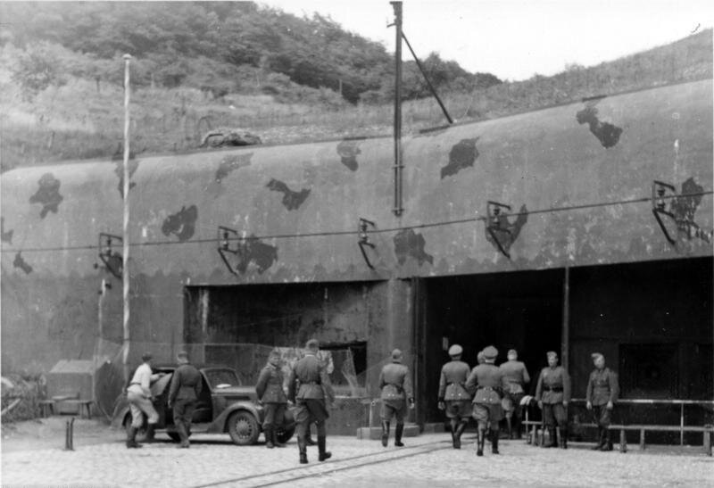 Na zdjęciu widać niemiecki bunkier. Niemieccy oficerowie i żołnierze kierują się w stronę wejścia. Bunkier częściowo jest wkopany w pagórek, jego zadaszenie pokrywa trwa. Wejście do bunkra jest wysokie i szerokie. U wejścia stoi samochód, do którego wsiada niemiecki żołnierz.