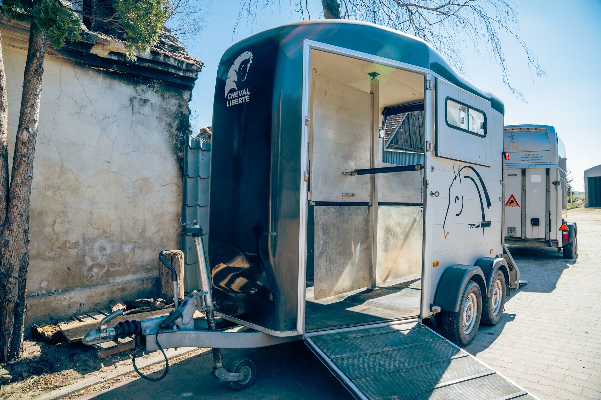 Zdjęcie przedstawia bukmankę, czyli przyczepę do transportu koni. Przyczepa posiada wejście z boku. Jest ono otwarte. Przed wejściem znajduje się rozłożona rampa. 