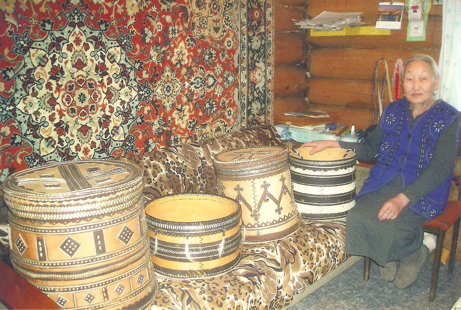  Na zdjęciu staruszka siedząca obok zdobionych, okrągłych pojemników. Na ścianie domu zawieszony jest dywan.