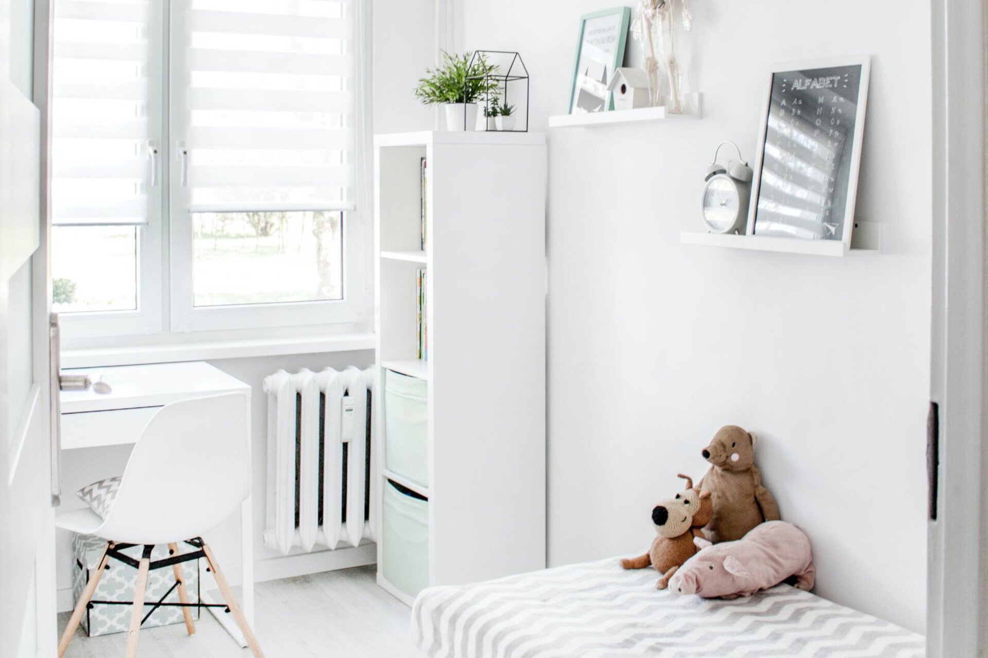 Zdjęcie przedstawia pokój dziecięcy, w którym od lewej strony znajdują się: biurko do pracy, krzesło, szafka, łóżko, na którym siedzą pluszowe zabawki. Nad łóżkiem znajdują się dwie półki ze stojącymi na nich obrazkami, domkiem, zegarem.