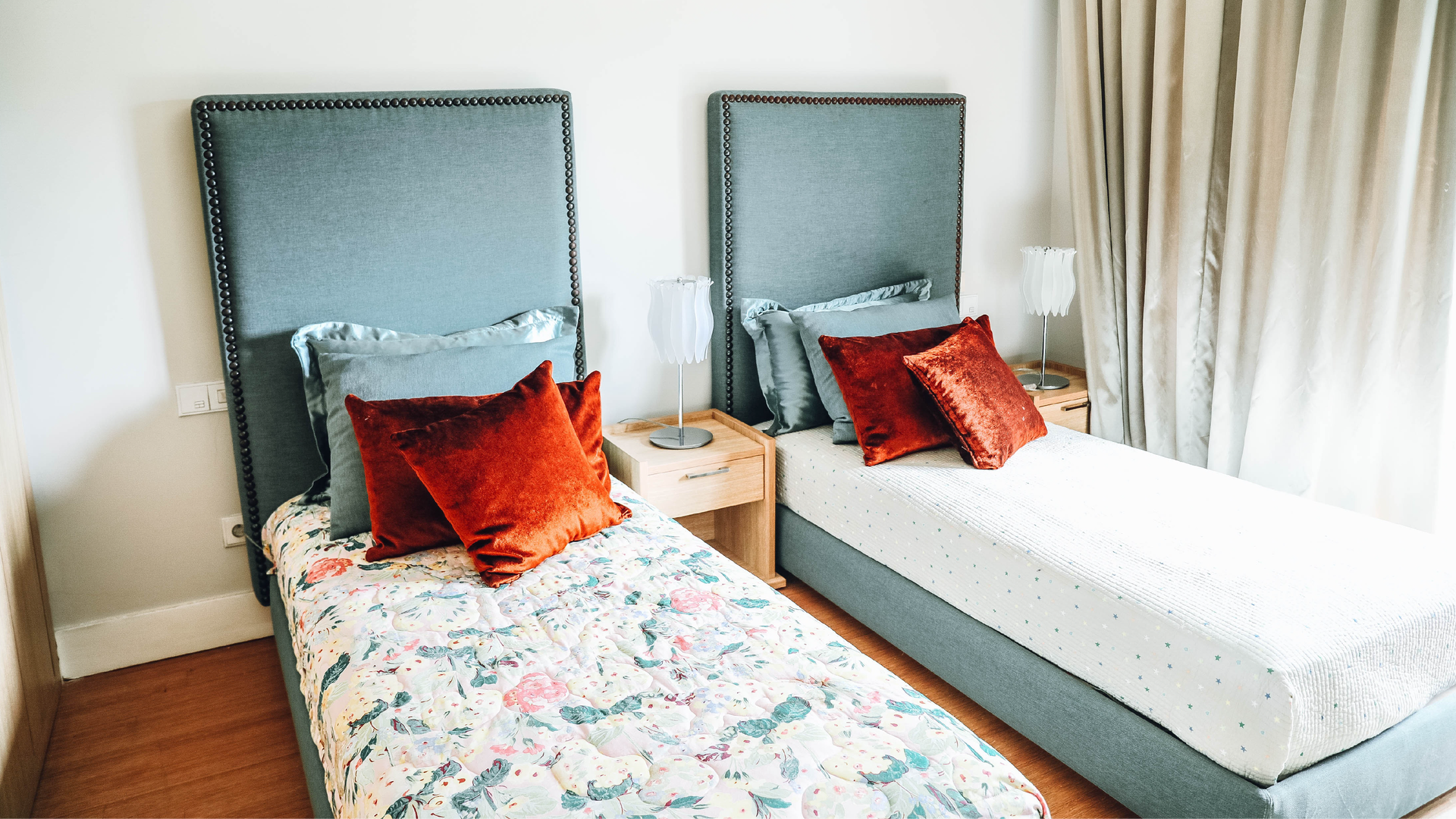 Zdjęcie przedstawia pokój hotelowy z dwoma pojedynczymi łóżkami. Każde z łóżek na inną narzutę. Na łóżkach widoczne są poduszki. Obok łóżek stoją małe stoliki nocne, a na nich małe lampy. Widoczna jest również zasłona.