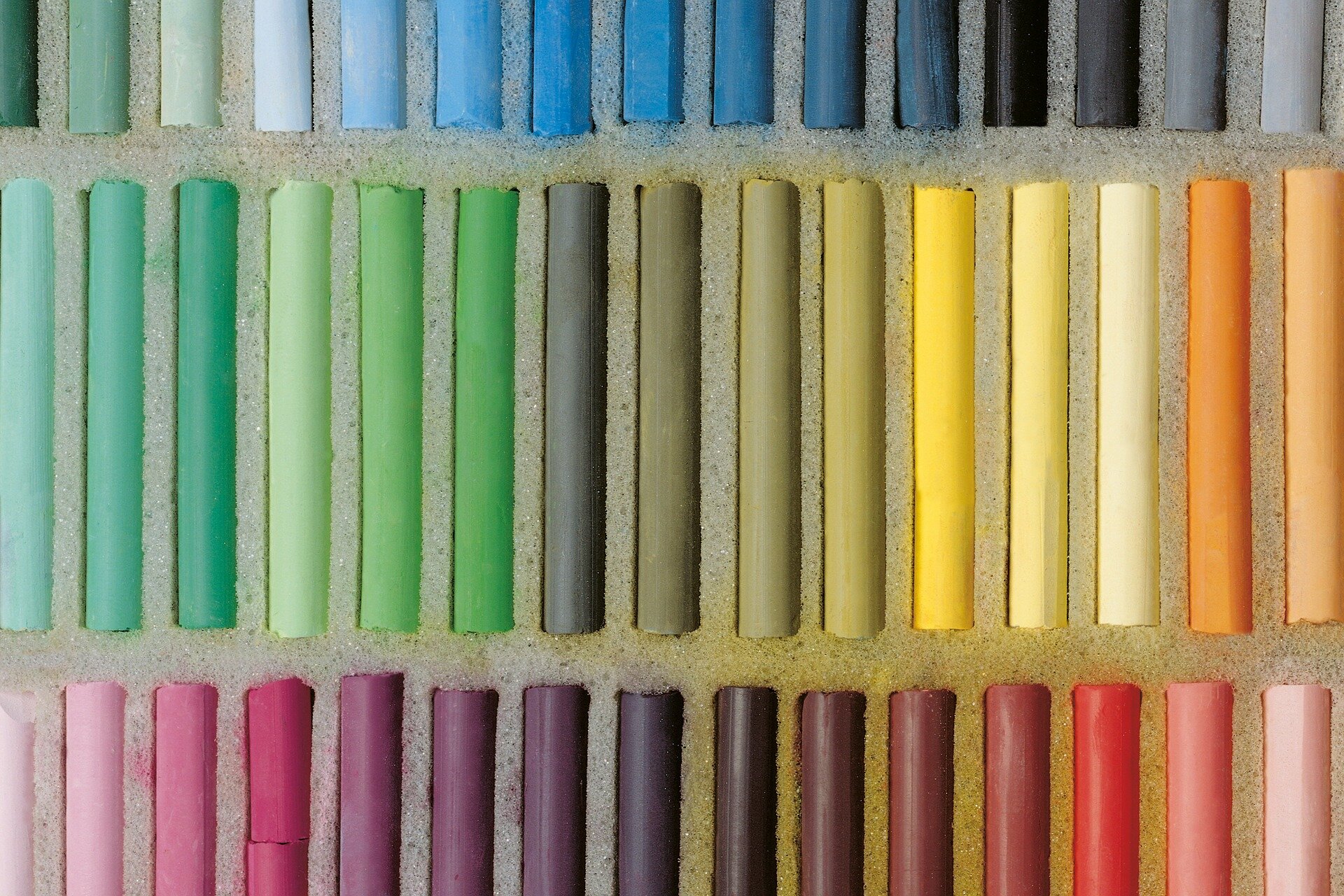 Zdjęcie przedstawia fragment zestawu kolorowych suchych pasteli. Kolorowe pałeczki o grubości centymetra i długości 8 centymetrów ułożone są równolegle do siebie w trzech poziomych rzędach. W górnym rzędzie przeważają odcienie błękitów i szarości, w środkowym od zieleni po lewej przechodzą przez oliwkowe na środku do żółtych i pomarańczowych po prawej. W dolnym rzędzie od jasnych fioletów z lewej, przez purpurę do brązów na środku po jaśniejące coraz bardziej czerwienie na prawo.