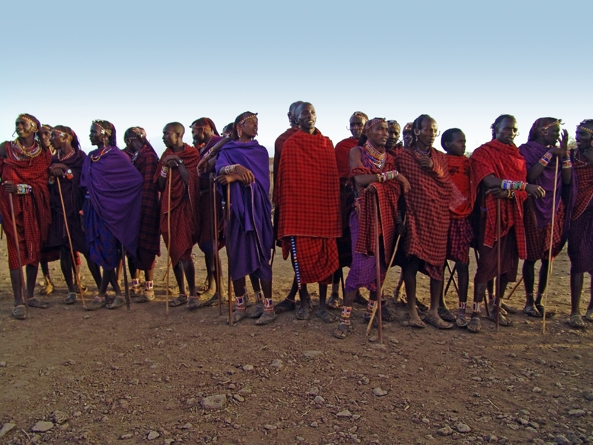 Fotografia przedstawia grupę masajskich wojowników. Mężczyźni mają ciemną skórę, są owinięci kolorowymi tkaninami – brązowymi, czerwonymi, w kratę. Wielu trzyma w rękach kije lub opiera się na nich. Mają pomalowane twarze i ozdoby na krótkich włosach. Na nogach sandały i opaski. Ludzie ci są uśmiechnięci, rozmawiają ze sobą.