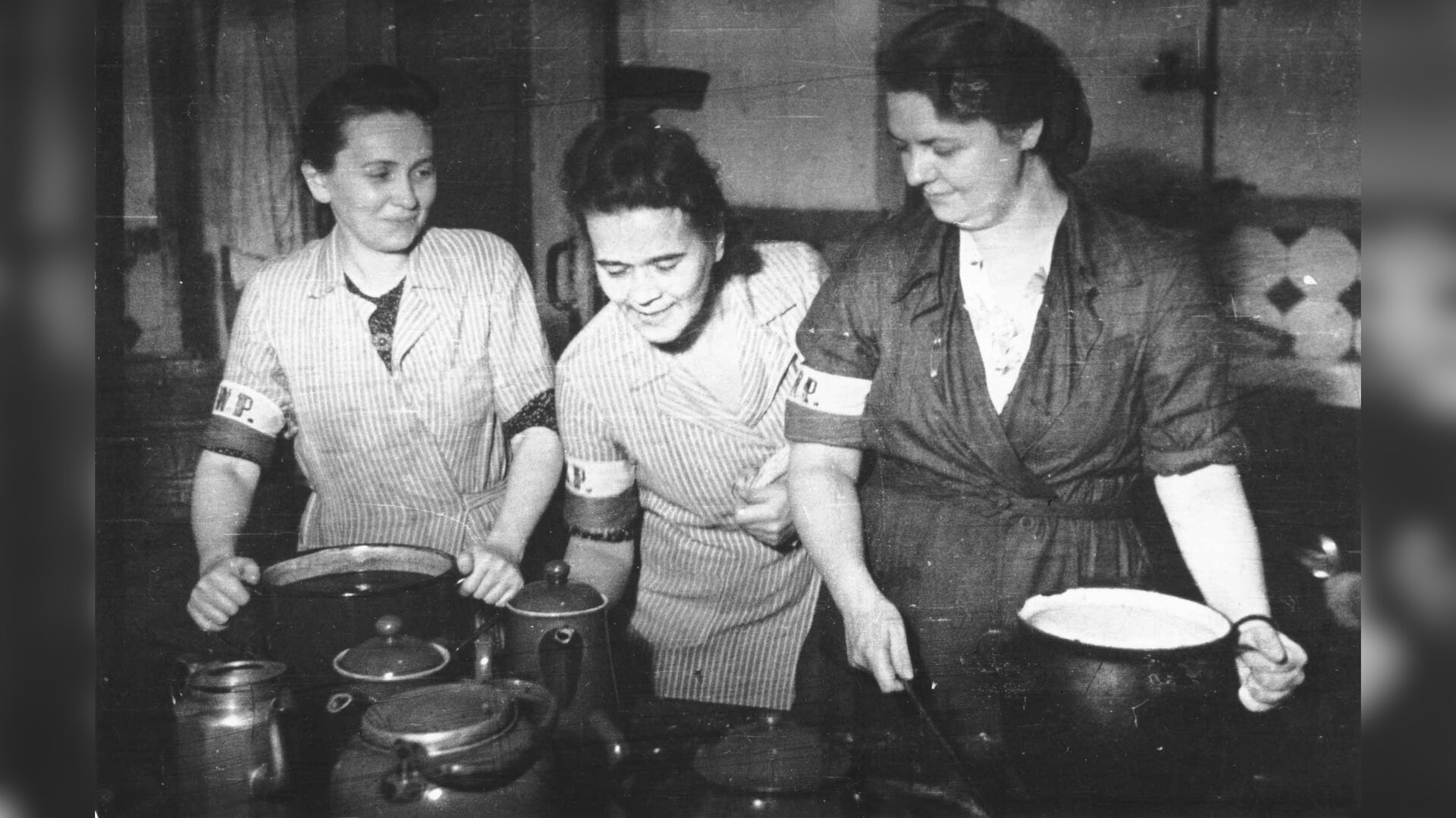 Zdjęcie przedstawia wnętrze pomieszczenia. Trzy kobiety w fartuchach z opaskami na ramieniu stoją przy garnkach stojących na kuchni.