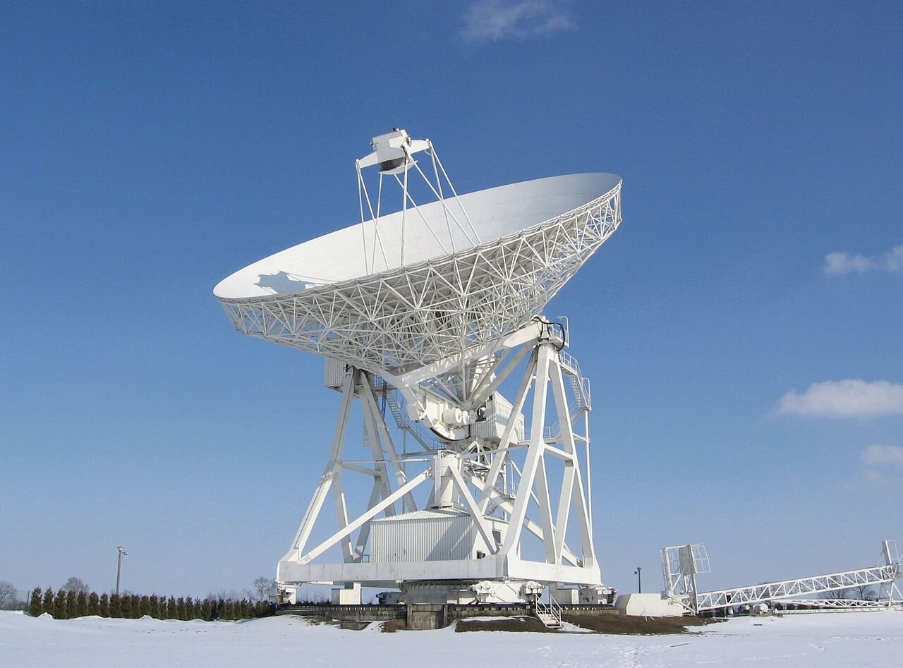 Zdjęcie przedstawia radioteleskop w miejscowości Piwnice pod Toruniem. Wielkie białe urządzenie ma kształt olbrzymiej misy stojącej na metalowej konstrukcji będącej stelażem.  Ze środka misy wystaje nadajnik na statywie. W tle błękitne, czyste niebo.