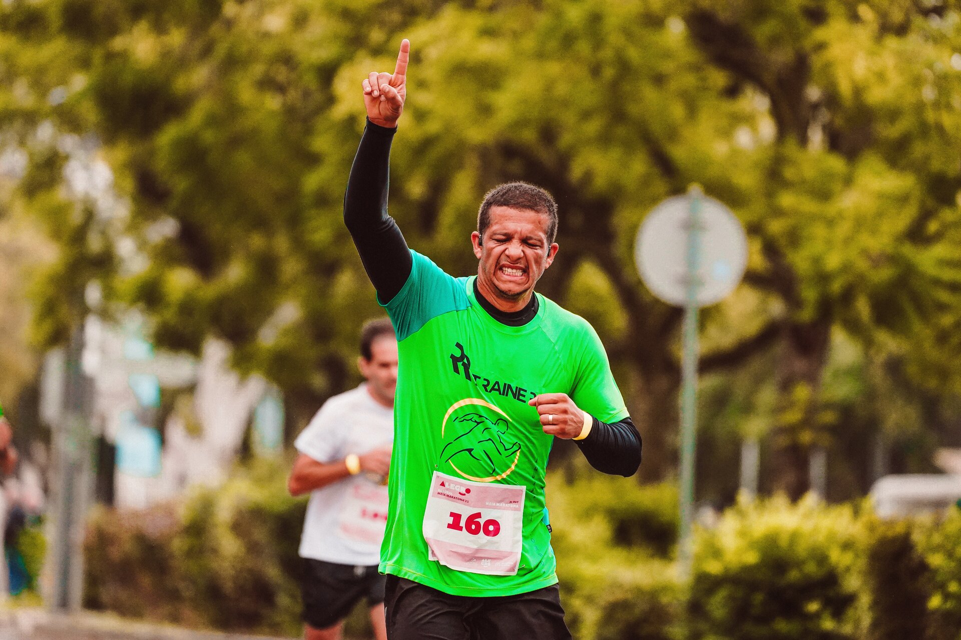 Zdjęcie przedstawia biegacza, w ujęciu od bioder w górę. Widoczny jest mężczyzna z krótkimi, ciemnymi włosami, ubrany w zieloną koszulkę z naklejonym numerem sto sześćdziesiąt. Jego prawa ręka jest podniesiona nad głowę, a palec wskazujący wysunięty i skierowany w niebo. Lewa ręka jest zgięta i przyciągnięta do tułowia. Na twarzy  mężczyzny widoczny jest grymas wysiłek. W tle za nim widoczna jest rozmyta postać drugiego biegacza oraz zielony żywopłot i drzewa.
