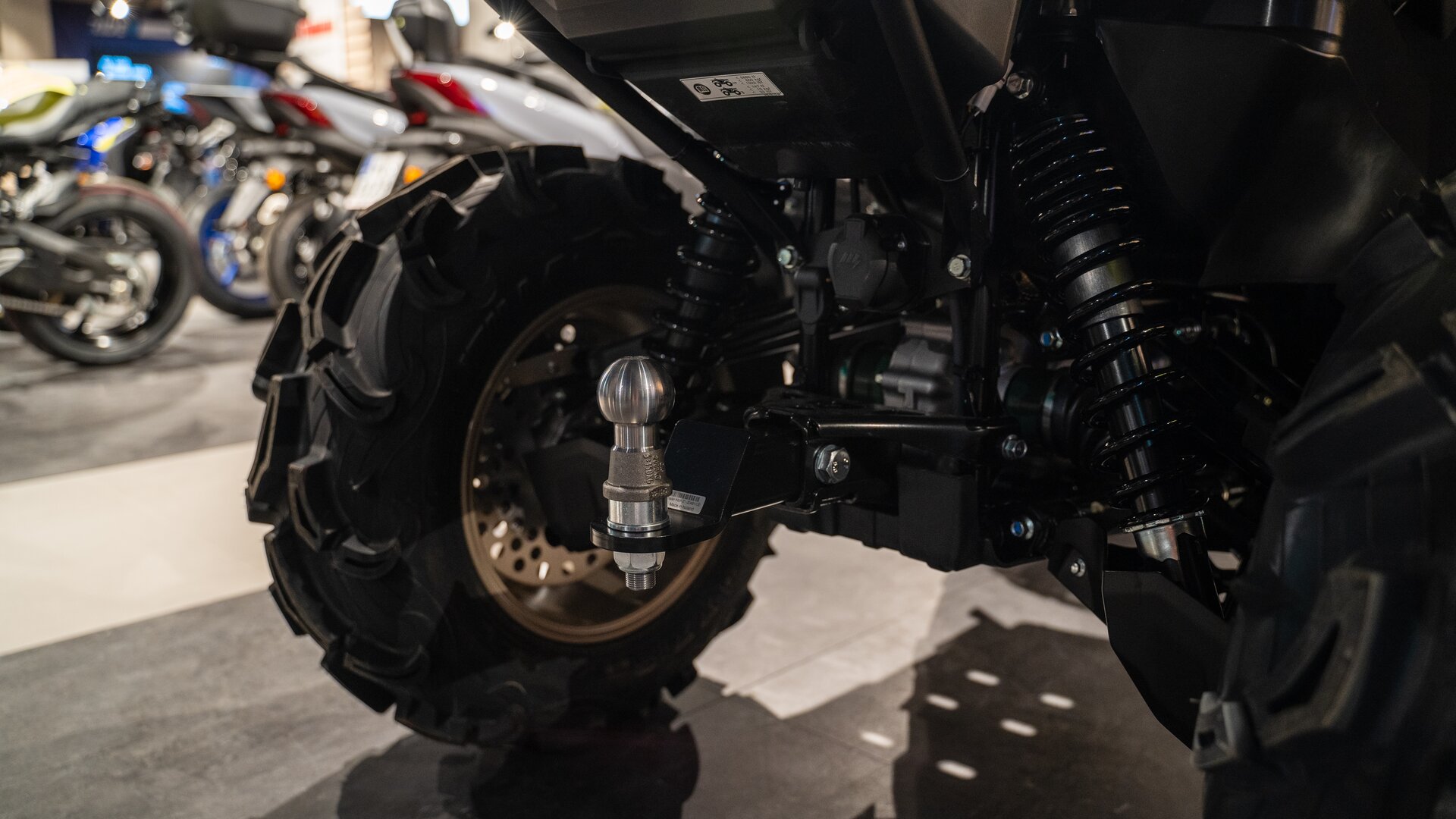 Zdjęcie ukazuje zbliżenie na tylne koło quada. Ma grubą oponę z wyraźnym bieżnikiem. Przy kołach widoczne amortyzatory, sprężyny oraz drążek do holowania. 