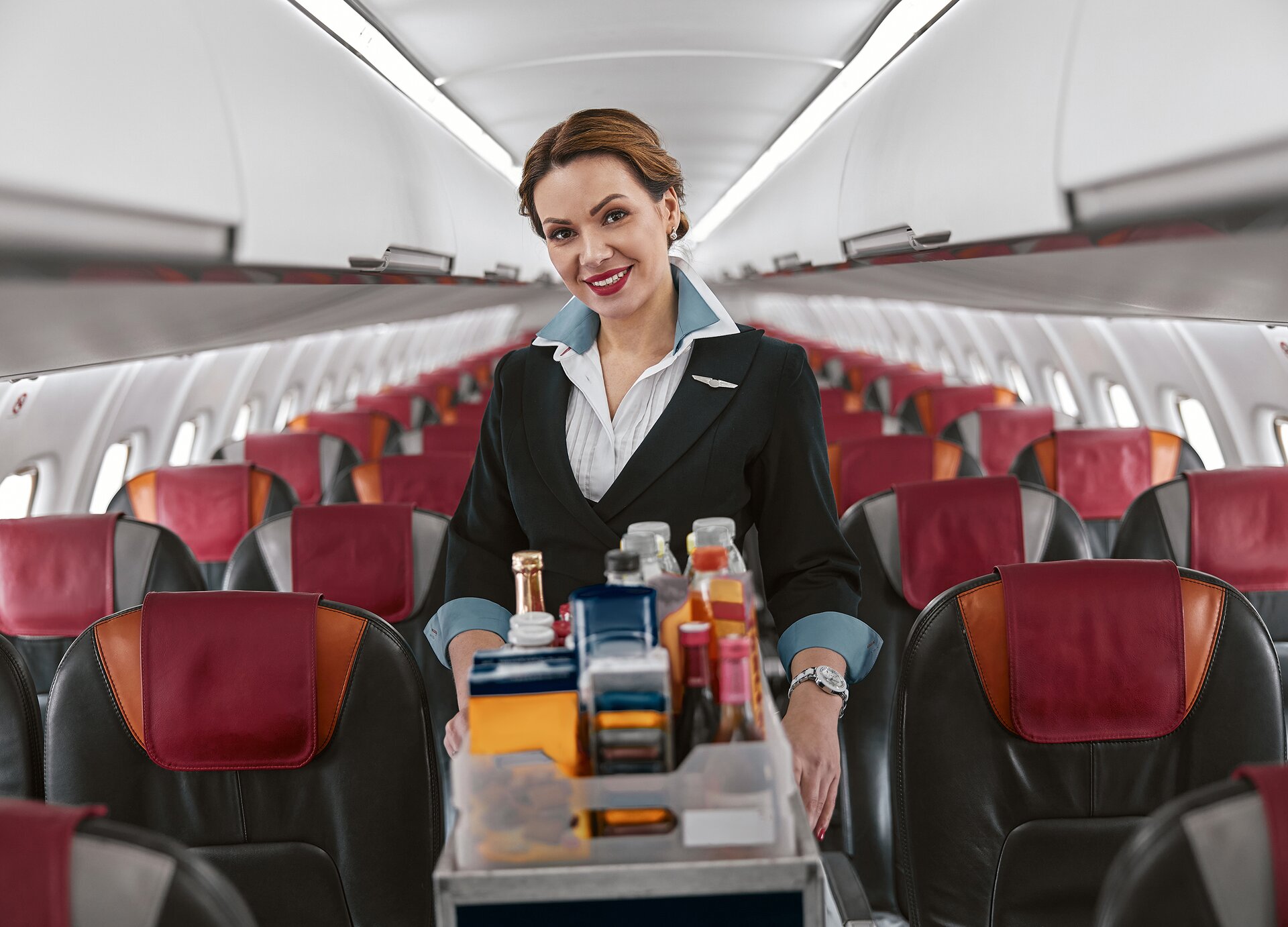 Zdjęcie przedstawia młodą kobietę, w ujęciu od pasa w górę, stojącą w przejściu w samolocie. Kobieta jest zwrócona w kierunku obiektywu, uśmiecha się. Jest ubrana w strój stewardessy, w ciemną marynarkę z szarymi mankietami, spod której wystaje biała koszula z dużym biało‑szarym kołnierzem. Obie dłonie ma oparte o wózek, na którym stoją różne napoje. Po prawej i lewej stronie widoczne są po dwa rzędy foteli pasażerskich.