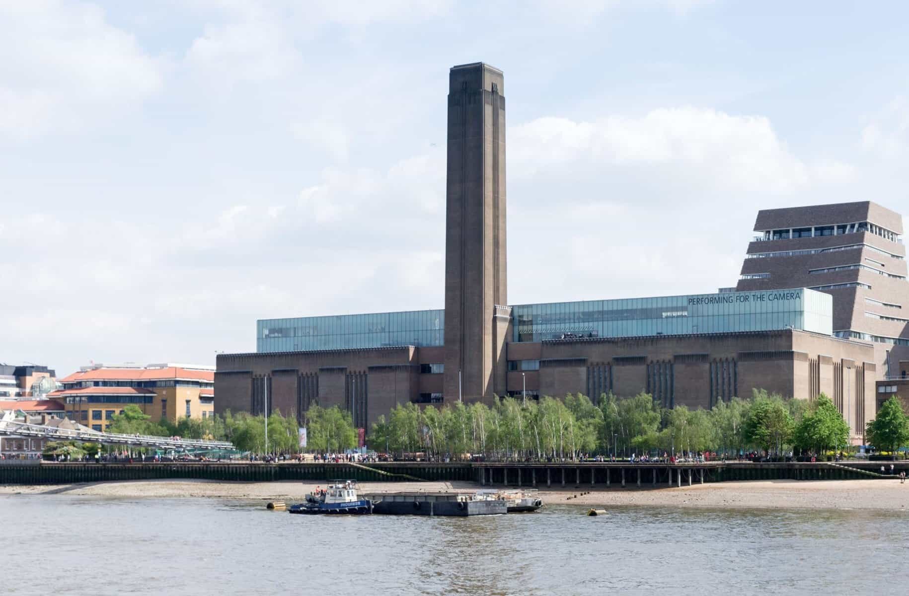 Fotografia przedstawia budynek o nieregularnym kształcie, znajdujący się nad wodą. Budynek ma niską, podłużną, murowaną bryłę, na której umieszczono przeszklony kawałek. Na środku budowli znajduje się bardzo wysoka murowana wieża o kształcie prostopadłościanu. z tyłu znajduje się część murowana ze szklanymi tarasami. Przed budynkiem rosną drzewa i stoją ludzie.