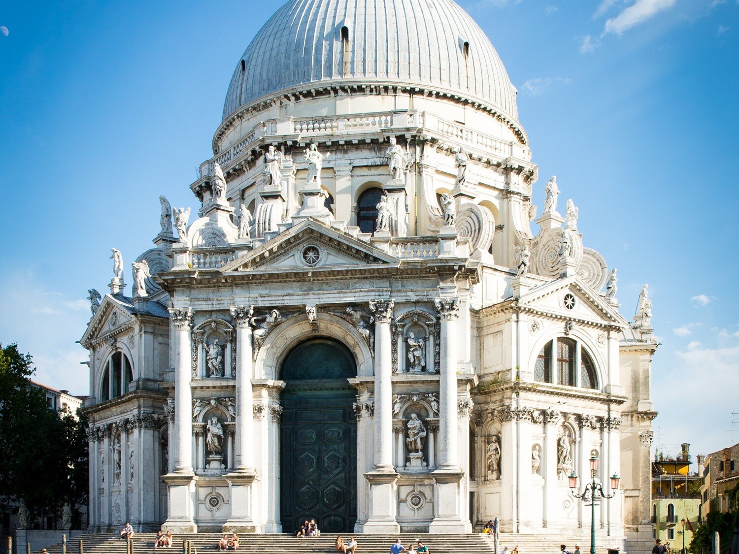 Zdjęcie przedstawia budynek świątyni zbudowany z białego kamienia na planie koła, z cylindrycznym elementem pośrodku zwieńczonym kopułą. Widoczna jest fasada z wysokim wejściem flankowanym dwoma parami  kolumn na wysokich postumentach, które zwieńczone są trójkątnym tympanonem. Po obu stronach wejścia widoczne są występy, które w górnej części mają półowalne okna, a w dolnej nisze z rzeźbami. Nad nimi, wokół cylindrycznej części, widoczne są rzeźby.