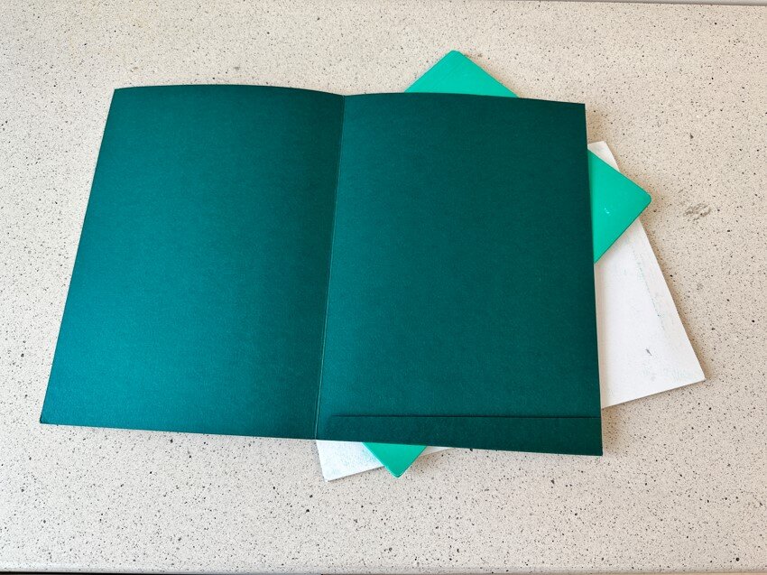 Zdjęcie przedstawia trzy teczki papierowej – w kolorach ciemnozielonym, jasnozielonym i białym. Ciemnozielona leży na wierzchu i jest rozłożona, tak że widać jej wnętrze.