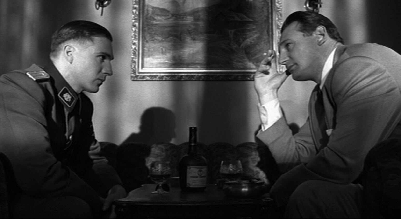 Zdjęcie przedstawia Schindlera siedzącego przy stoliku. Naprzeciw niego siedzi niemiecki oficer w mundurze. Mężczyźni piją alkohol.