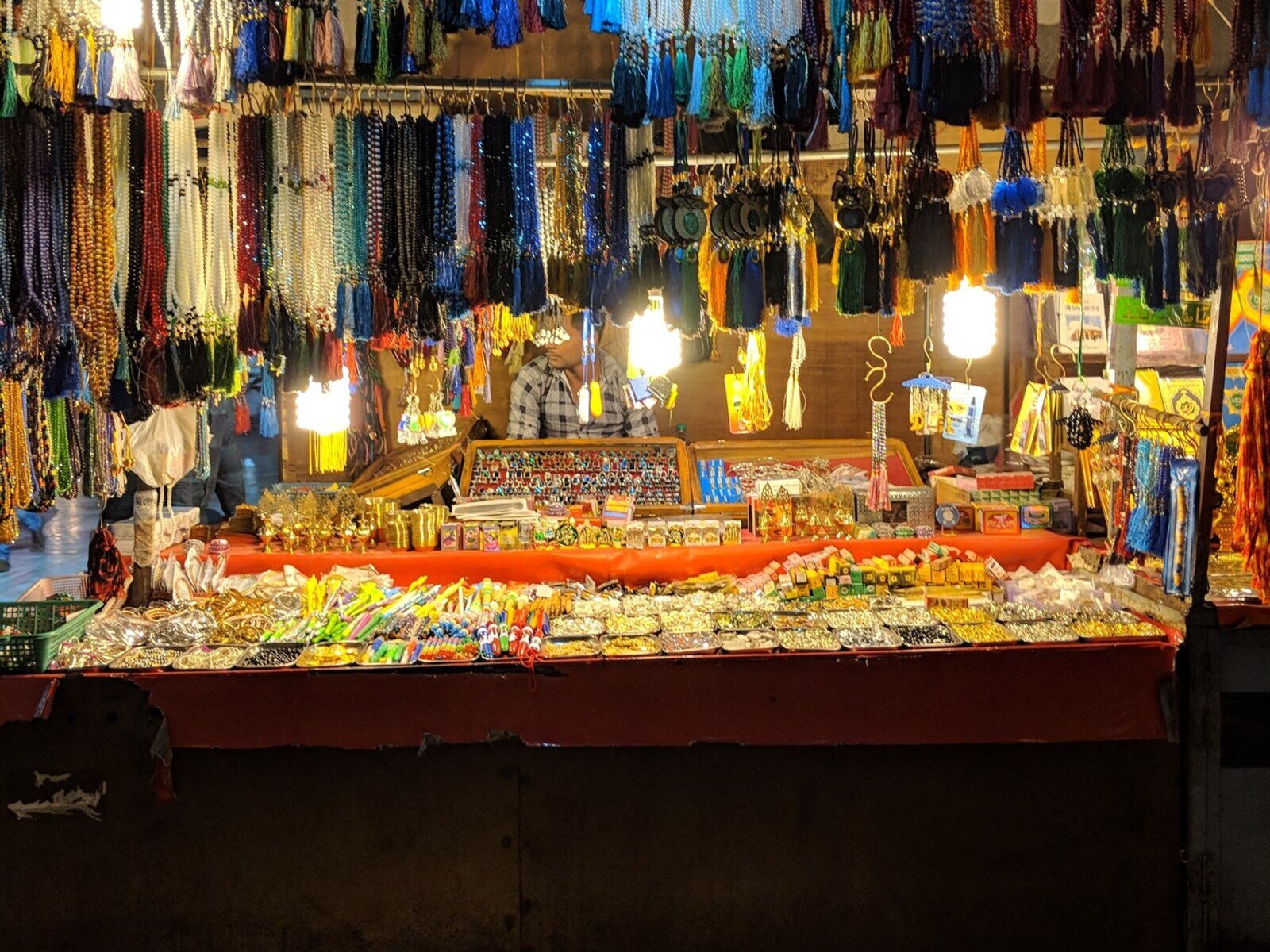 Zdjęcie przedstawia kolorowe stoisko na bazarze, na którym wystawione są korale, pierścionki, upominki, drobne zabawki, kadzidła, zawieszki, bransoletki, a także drobne złote pamiątki. W tle, za sznurem wiszących ozdób, widoczna jest postać siedzącego mężczyzny, zwróconego w lewo. Stoisko jest podświetlone trzema lampami. 
