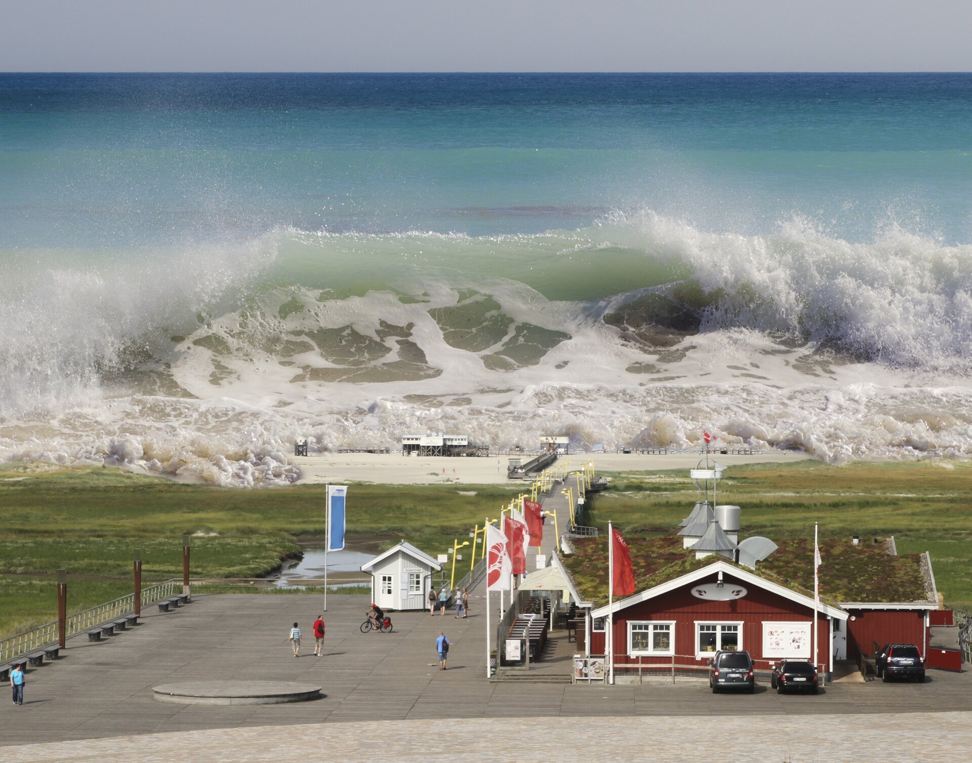 Zdjęcie przedstawia ogromną falę tsunami zbliżającą się do brzegu, na którym znajduje się kilka parterowych budynków i zaparkowane samochody.