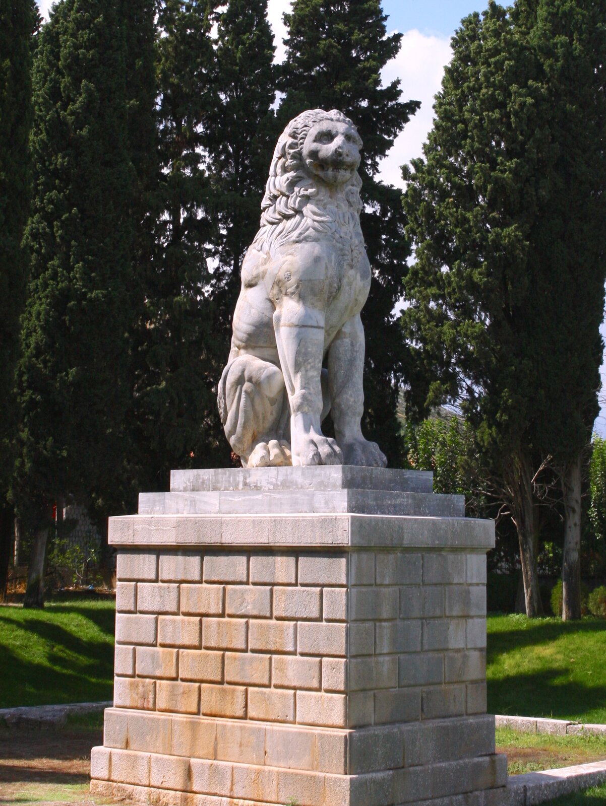 Zdjęcie przedstawia rzeźbę lwa, która stoi na postumencie. W tle rosną wysokie drzewa.