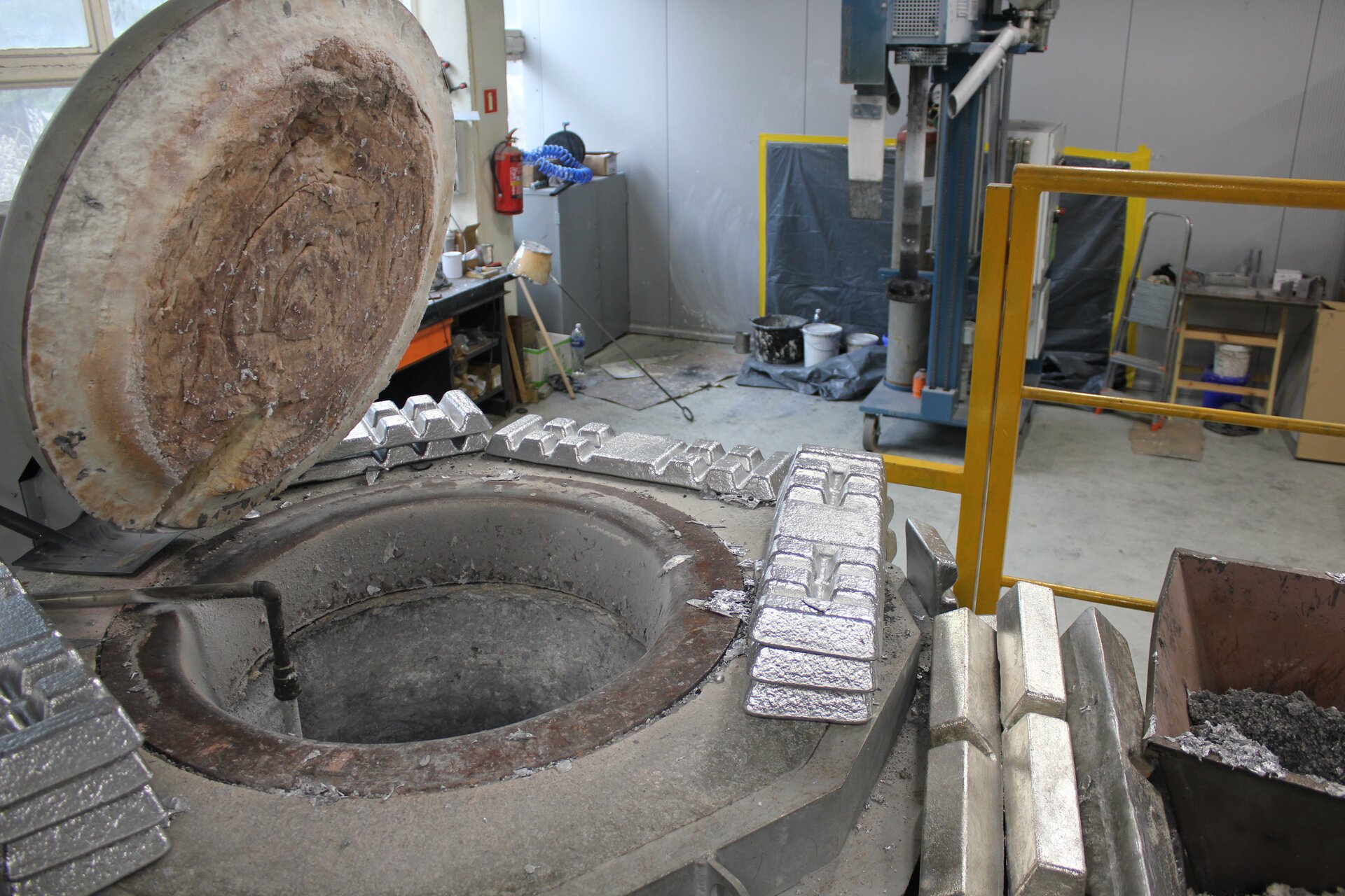 Ilustracja przedstawia otwarty piec odlewniczy z wymurówką. Wokół pieca ułożone są gąski metalowe. Wewnątrz pieca oraz na tyglu, widoczna jest gruba warstwa wymurówki.