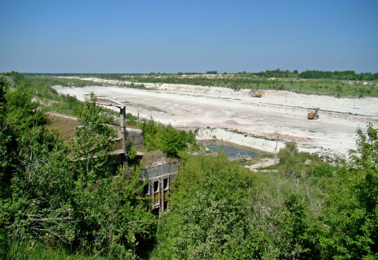 Zdjęcie przedstawia kopalnie odkrywkową. Na pierwszym planie zielona roślinność. W gąszczu drzew widoczny betonowy opuszczony budynek. W tle ogromny plac piasku, na placu dwie maszyny przemysłowe, W tle ściany kopalni porośnięte zieloną roślinnością.