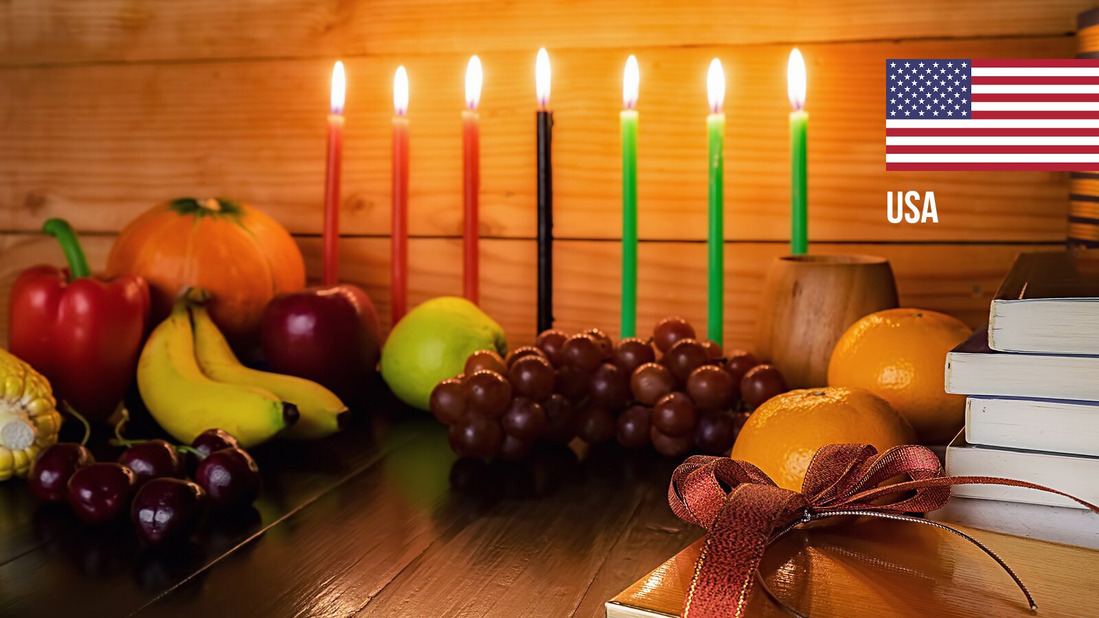 Zdjęcie przedstawia drewniany blat, na którym – na tle drewnianej ściany – widać siedem cienkich zapalonych świec. Od lewej trzy świece czerwone, jedna czarna i trzy zielone. Przed świecami oraz po lewej stronie widoczne są ułożone owoce i warzywa, takie jak: fioletowe winogrona, banany, czerwona papryka, fragment kolby kukurydzy, czereśnie, jabłko, gruszka, dynia. Po lewej stronie widoczne są pomarańcze, książki ułożone w stos, drewniany kubek i fragment pudełka prezentowego z kokardą. W prawym górnym rogu widoczna jest flaga USA.