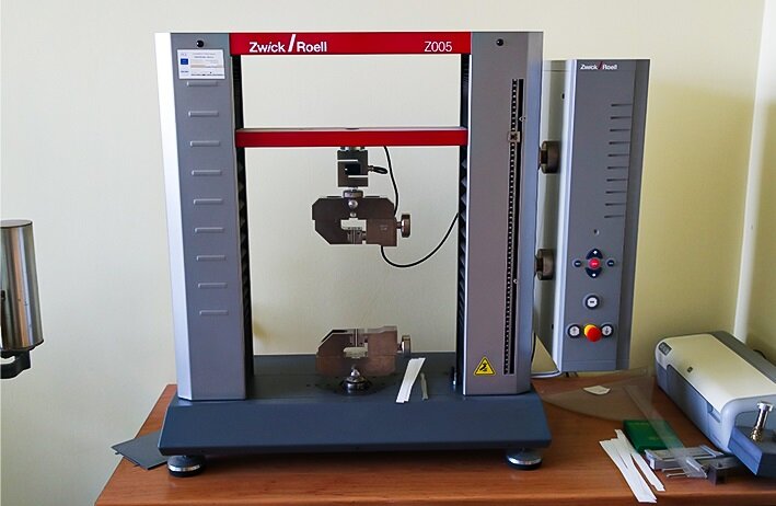 Zdjęcie przedstawia zrywarkę. Widoczny jest metalowy stelaż urządzenia złożony z poziomej podstawy na niskich, okrągłych nóżkach. Z prawej i lewej strony podstawy wychodzą dwa metalowe, pionowe przęsła, połączone w górnej części dwiema poprzeczkami w kolorze czerwonym. Do prawego przęsła przymocowany jest prostopadłościenny, przesuwany w górę i w dół, element, w którego dolnej części widoczne są przyciski i pokrętła. Do czerwonej poprzeczki, umieszczonej  na wysokości 3/4 przęseł, pośrodku przymocowany jest element z pokrętłem, który znajduje się dokładnie nad zaciskiem pośrodku podstawy. Na prawo od zrywarki widoczne są paski papieru, przeźroczysta linijka, drukarka.