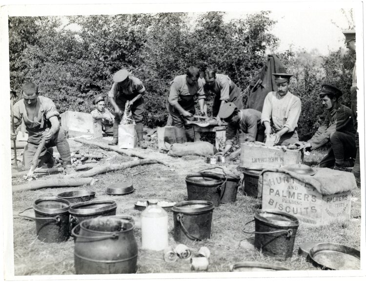 Zdjęcie przedstawia grupę żołnierzy - kucharzy przygotowujących jedzenie. Na pierwszym planie widać duże garnki, wiadra, baniaki. Na drugim planie, żołnierze krojący i przygotowujący jedzenie na skrzyniach z produktami, po lewej stronie mężczyzna układający drewno na palenisko.
